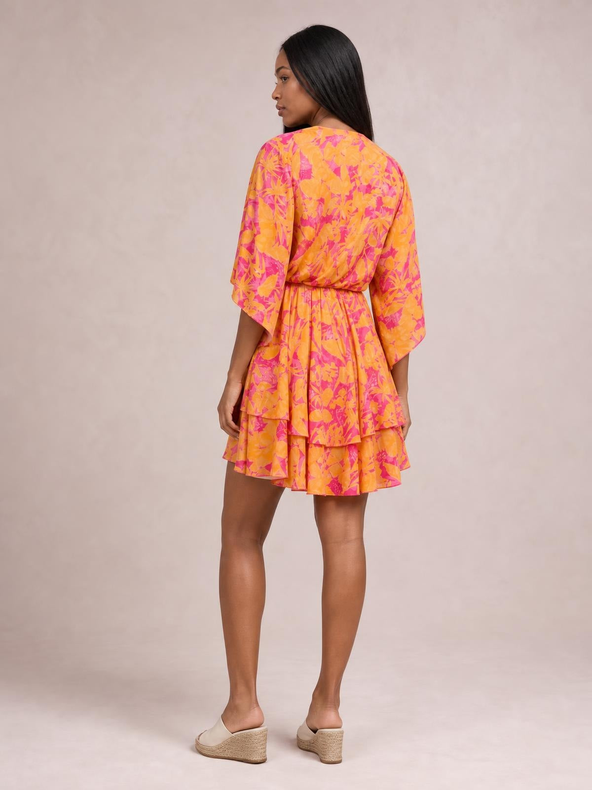 Model poses in a three-quarter back view wearing a bright orange and pink printed mini dress with long sleeves, cinched waist, and layered tiered skirt.