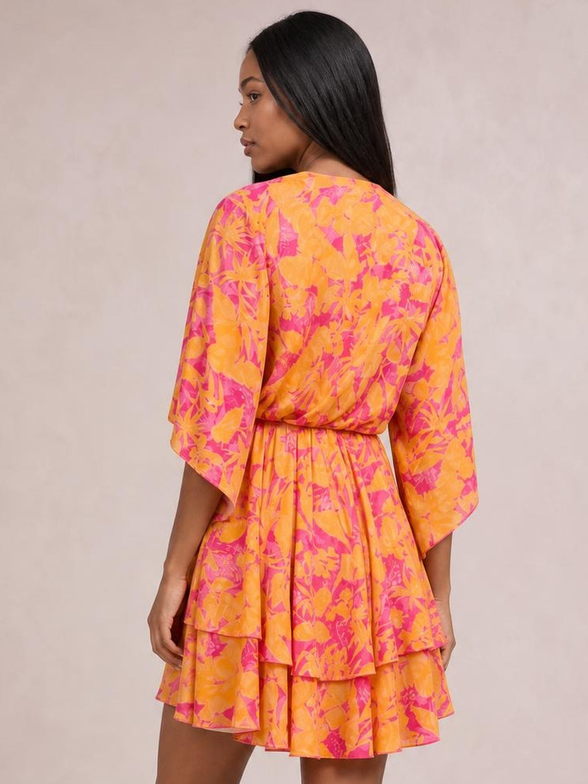 Model looks over her shoulder in a bright orange and pink printed mini dress with a belted waist, ruched detailing, flared sleeves, and tiered ruffle layers.