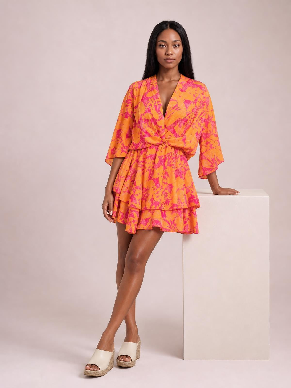 A model poses in a bright orange and pink printed surplice mini dress with a ruched tie waist, tiered ruffle skirt, and three-quarter sleeves.