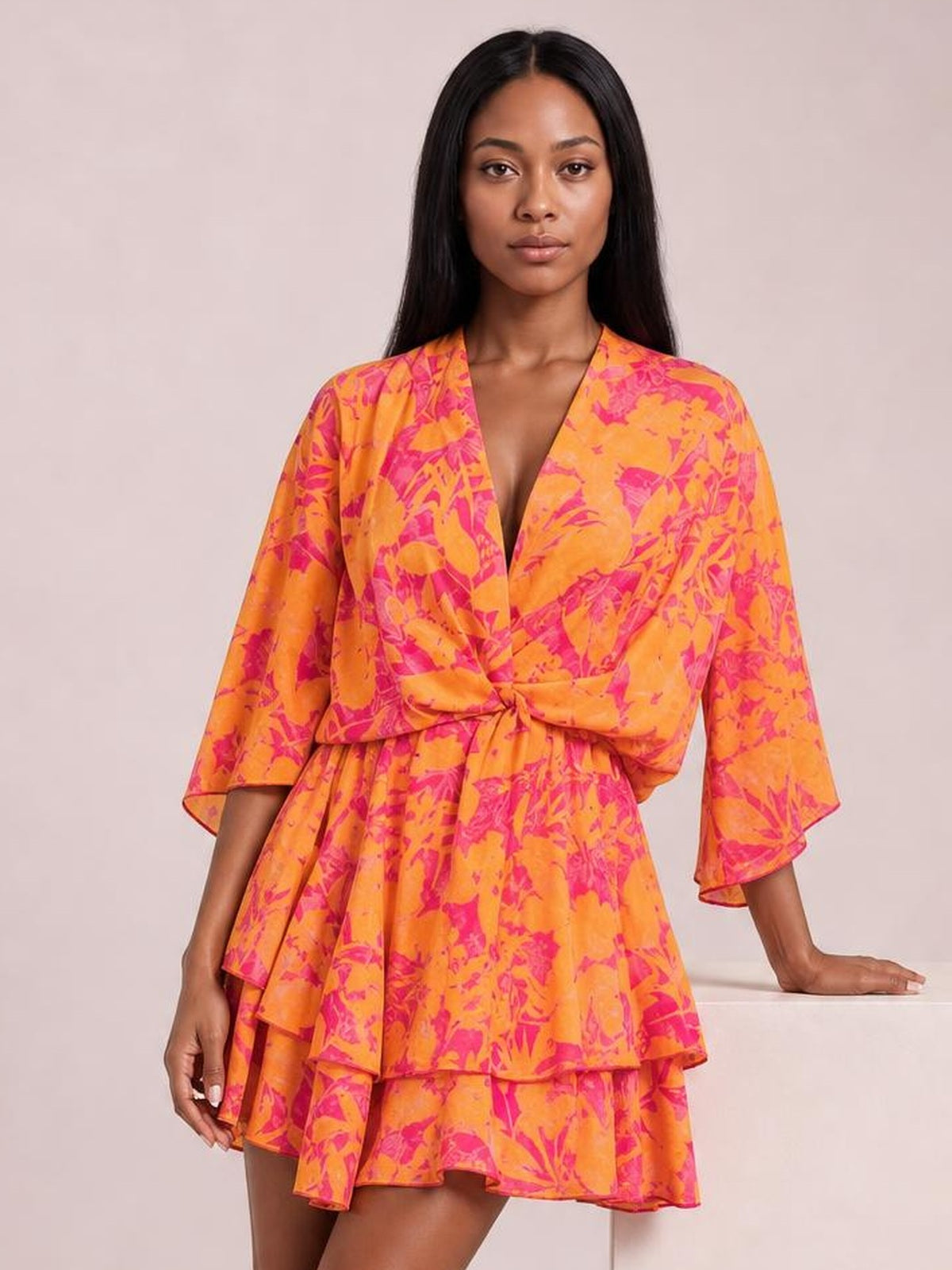 Model poses in a bright orange and pink printed surplice mini dress with long sleeves, a tied waist, gathered details, and tiered ruffled layers.