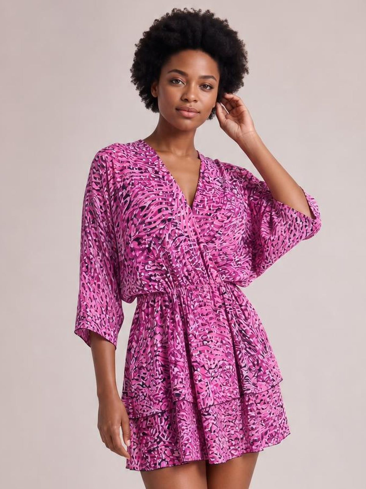A model poses in a pink and purple printed surplice mini dress with a V-neckline, belted ruched waist, tiered skirt, and flowing three-quarter sleeves.