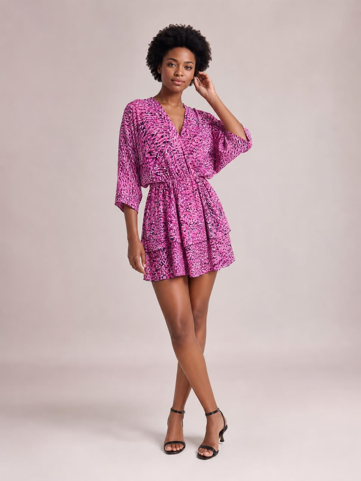 A model poses with crossed legs in a pink and purple printed surplice mini dress featuring a V-neck, ruched belted waist, tiered skirt, and loose sleeves.