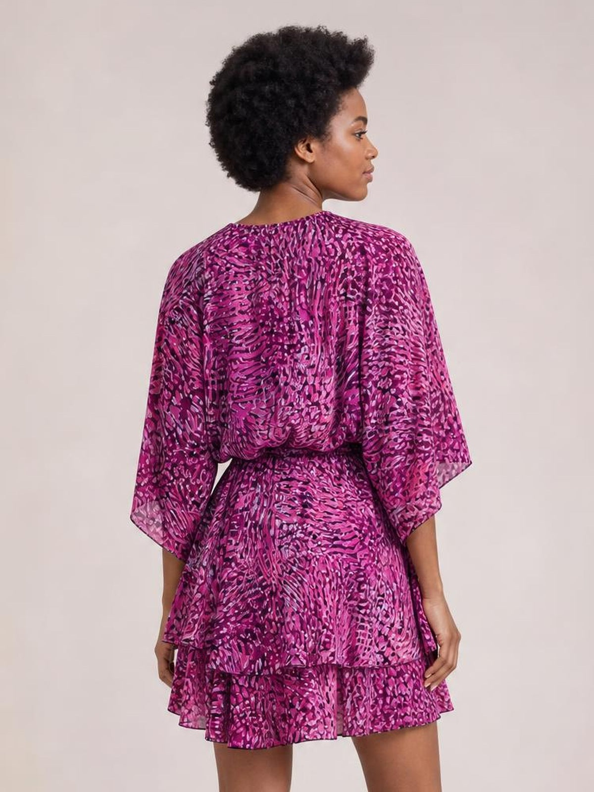 A model shows the back of a purple printed surplice mini dress with voluminous sleeves, a belted waist, tiered construction, and a flared ruffled hem.