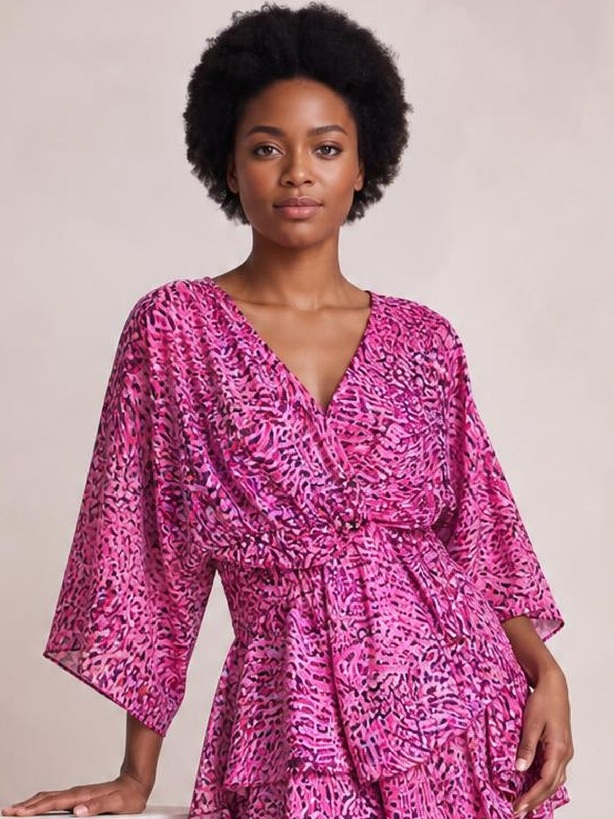Model poses in a vibrant pink printed surplice mini dress with ruched waist, tiered silhouette, and flared three-quarter sleeves.