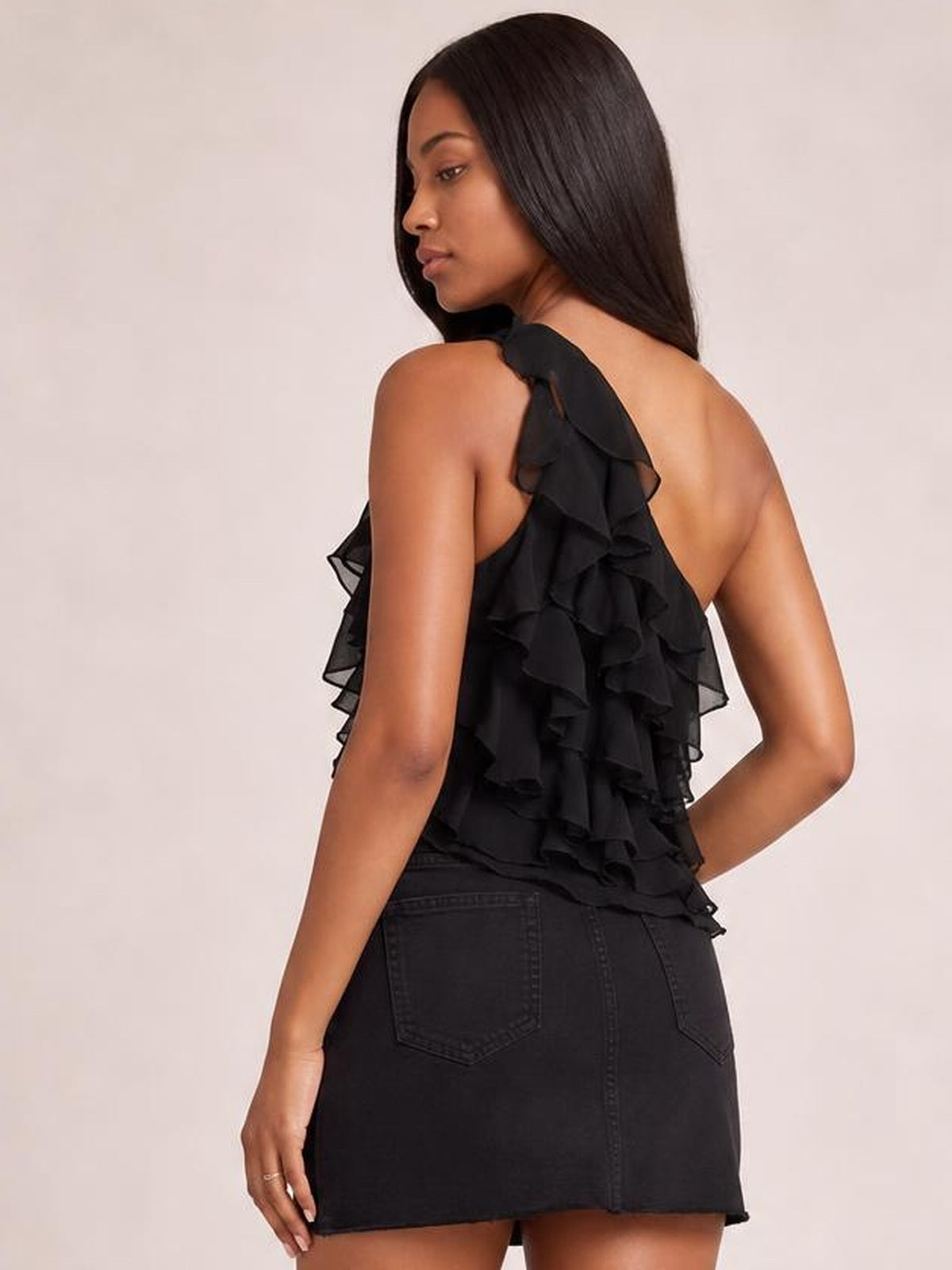 Model turns to show a black one-shoulder tank with cascading layered ruffles, an asymmetrical silhouette, and an open back.