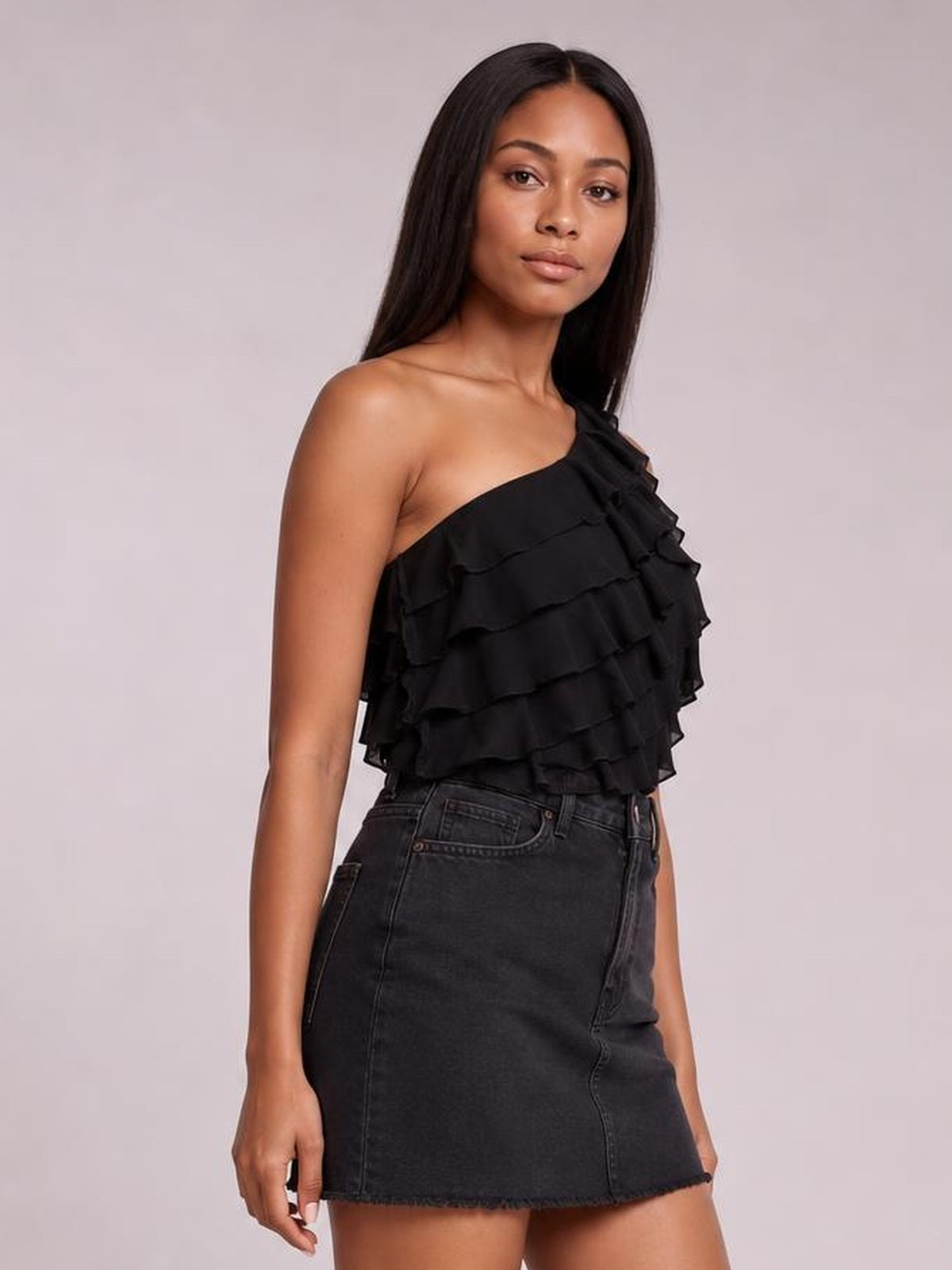 Model poses in a black layered ruffled one-shoulder tank with a cropped, voluminous silhouette and asymmetrical neckline, paired with a dark denim skirt.