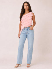 A model stands with one hand in her jeans pocket wearing a pink one-shoulder tank with layered ruffles and an asymmetrical cropped silhouette.