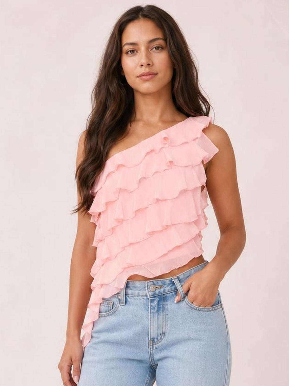 A model poses in a pink layered ruffled one-shoulder tank with an asymmetrical neckline, flowing silhouette, and cascading tiered construction.