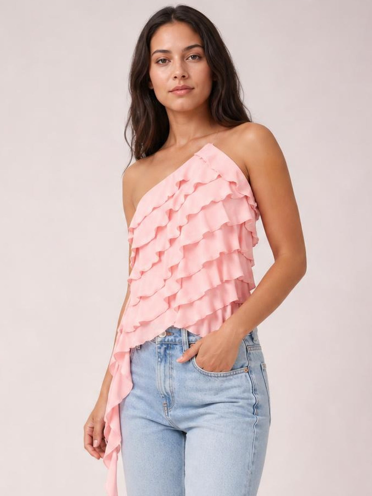 Model in a relaxed pose wearing a pink layered ruffled one-shoulder tank with an asymmetrical draped hem and light-wash jeans.