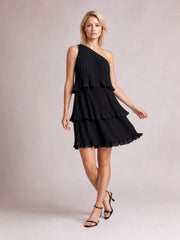 A model poses with crossed legs in a black layered one-shoulder mini dress featuring fine pleats, tiered ruffled panels, and a softly flared silhouette.