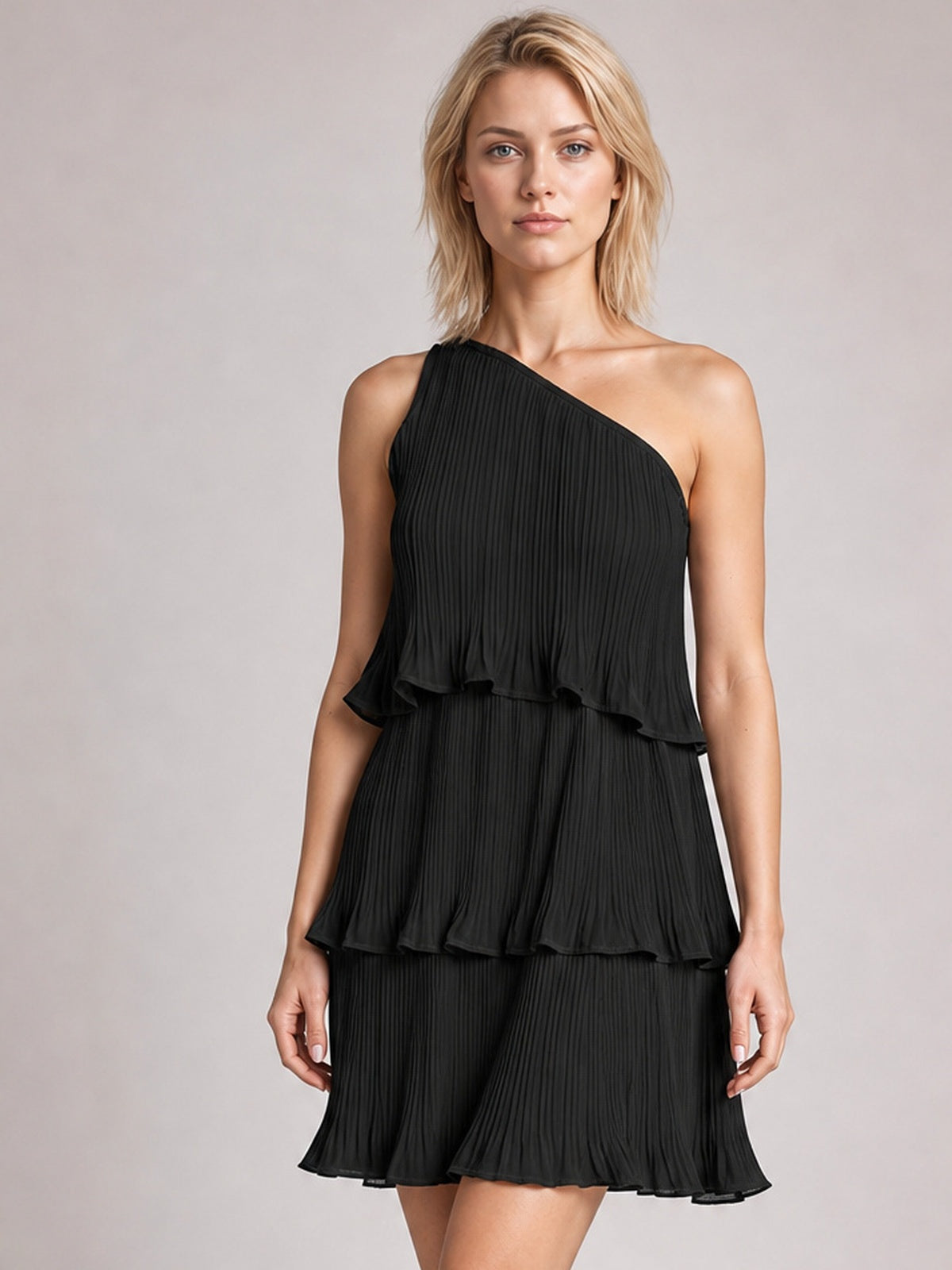 A model stands in a black single-shoulder mini dress with fine pleats, layered ruffled tiers, an asymmetrical neckline, and a flowing A-line silhouette.