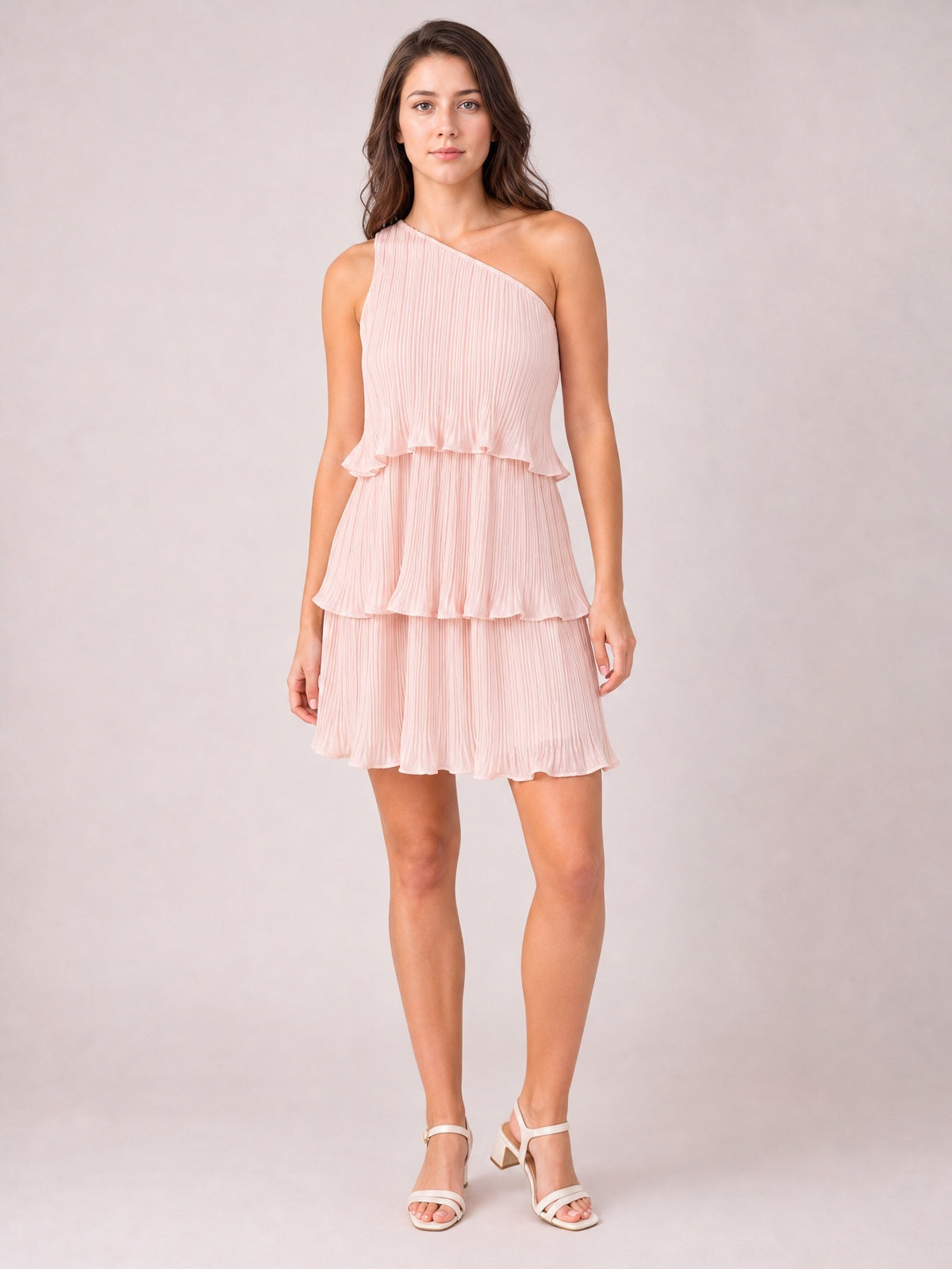 Model stands in a blush pink single-shoulder mini dress with fine pleats, ruffled layered tiers, an asymmetrical neckline, and a softly flared silhouette.