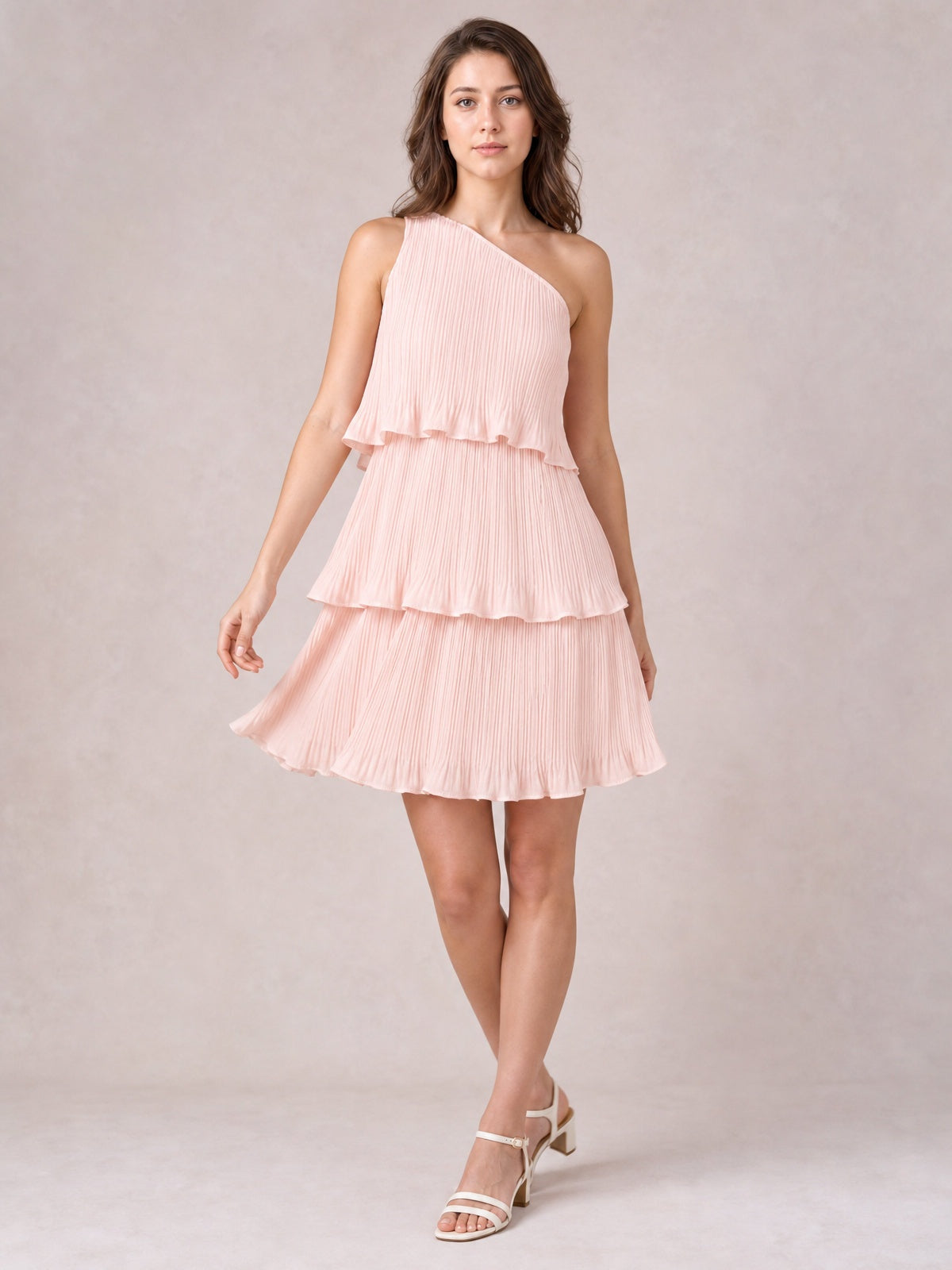 Model poses in a pale pink layered mini dress with a single-shoulder neckline, ruffled tiers, fine pleats, and a softly flared silhouette.