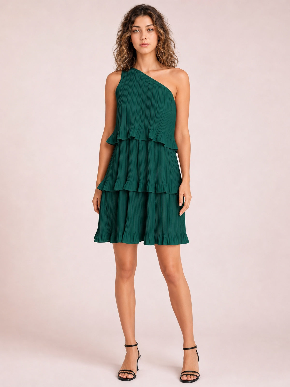 A model stands in a deep green single-shoulder mini dress with fine pleats, layered ruffled tiers, an asymmetric neckline, and a softly flared silhouette.