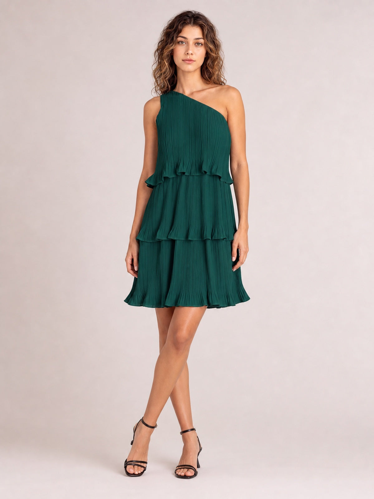 Model poses with crossed legs in a dark green single-shoulder mini dress featuring a pleated texture, ruffled layers, and a tiered flared silhouette.