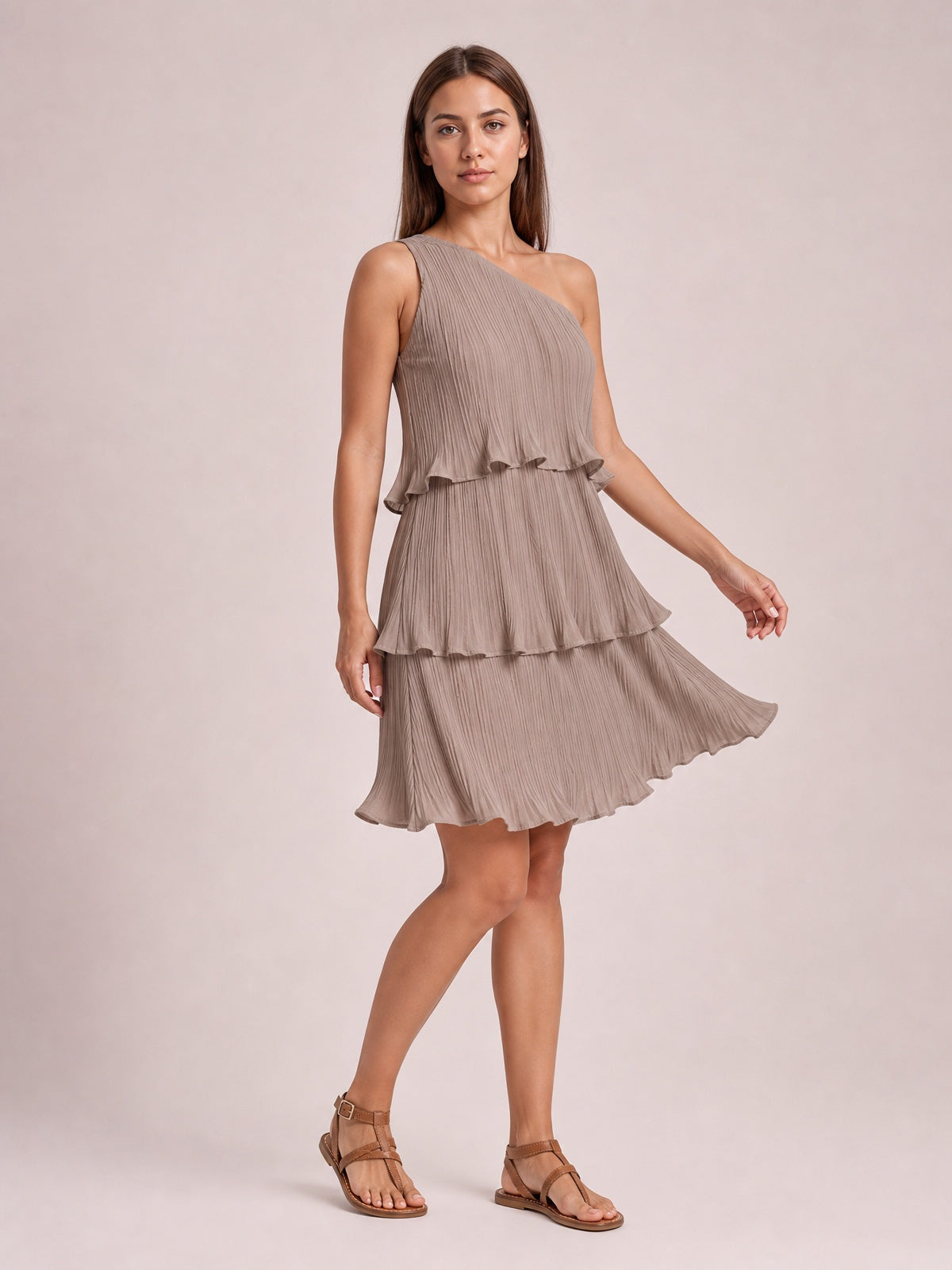 Model poses in a taupe single-shoulder mini dress with a pleated texture, layered ruffled tiers, and a softly flared silhouette.