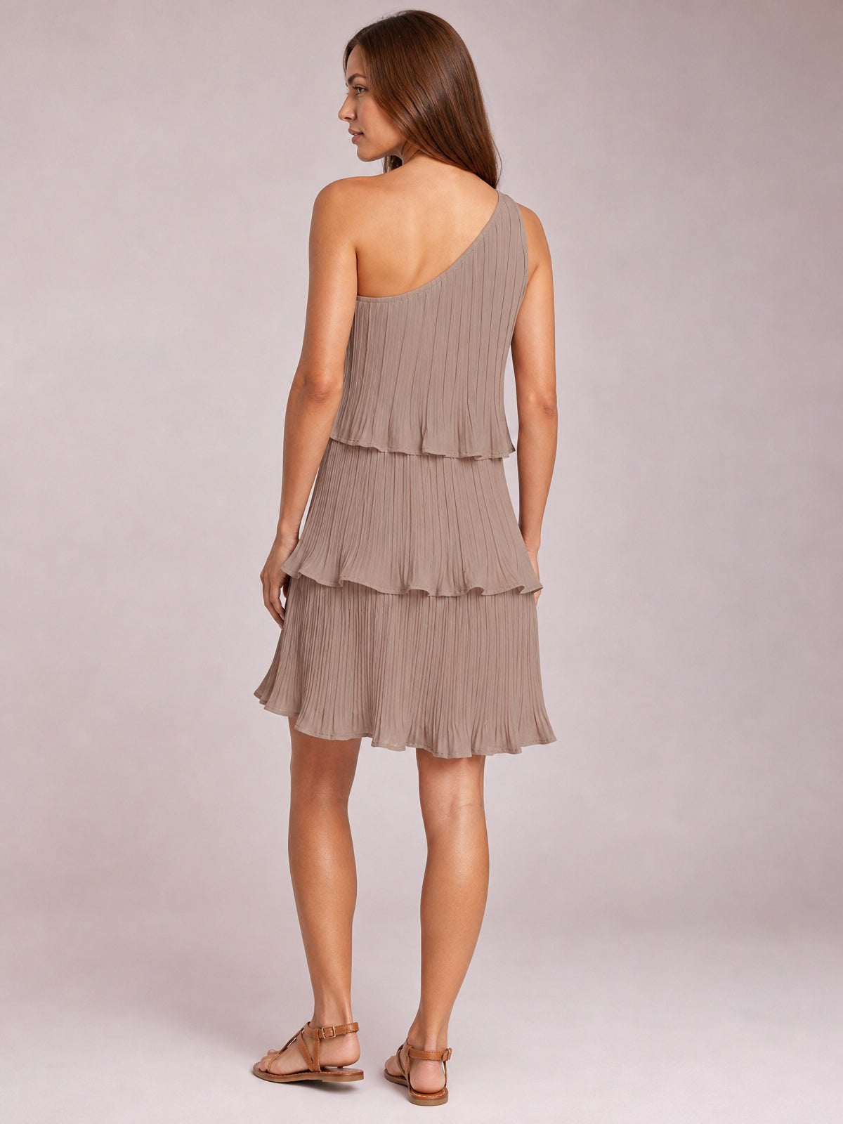 Model shown from behind in a taupe layered single-shoulder mini dress with fine pleats, tiered ruffled panels, and a softly flared silhouette.