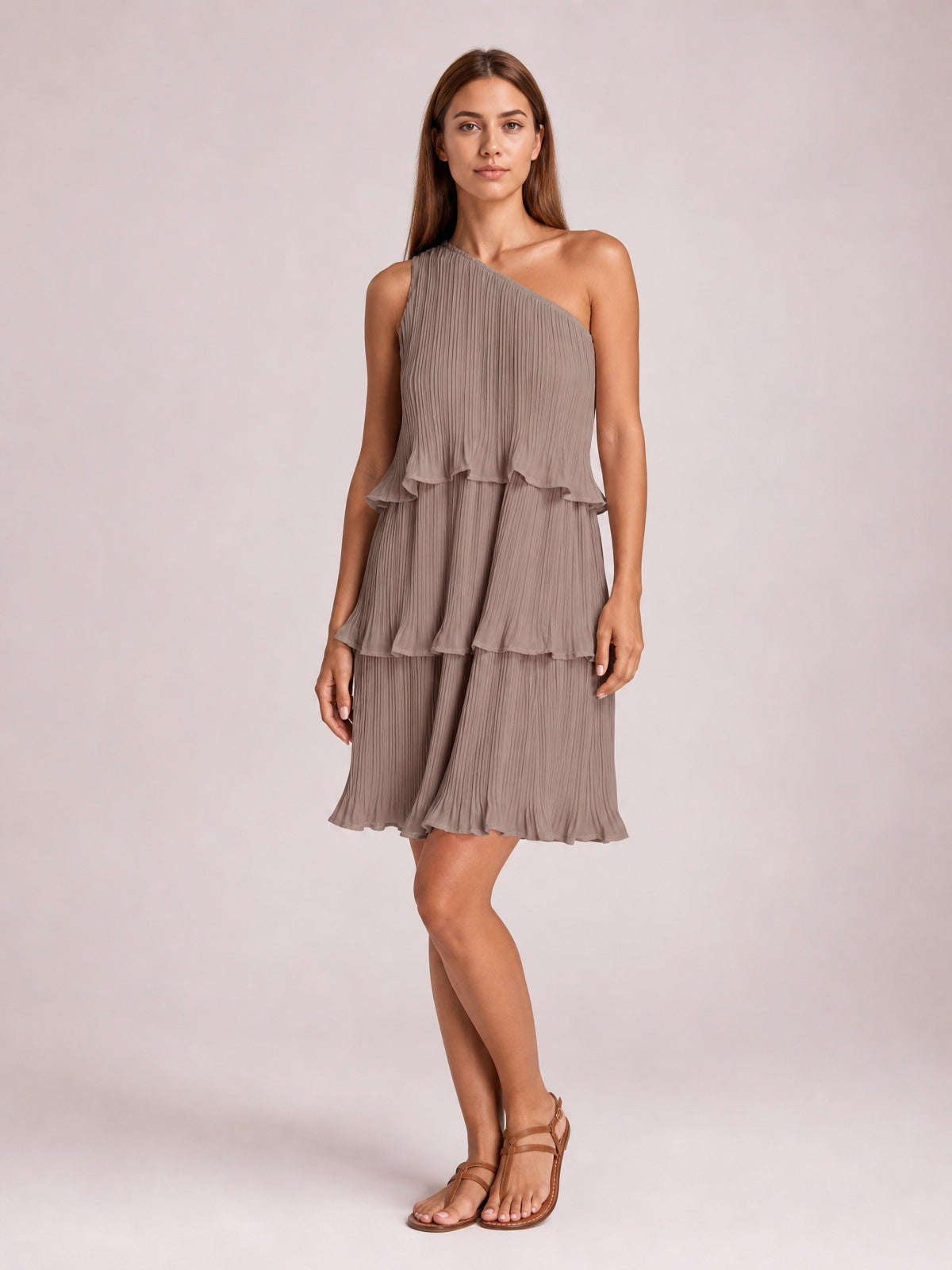 A model stands with crossed legs in a taupe single-shoulder mini dress featuring fine pleats, layered ruffled tiers, and a relaxed silhouette.