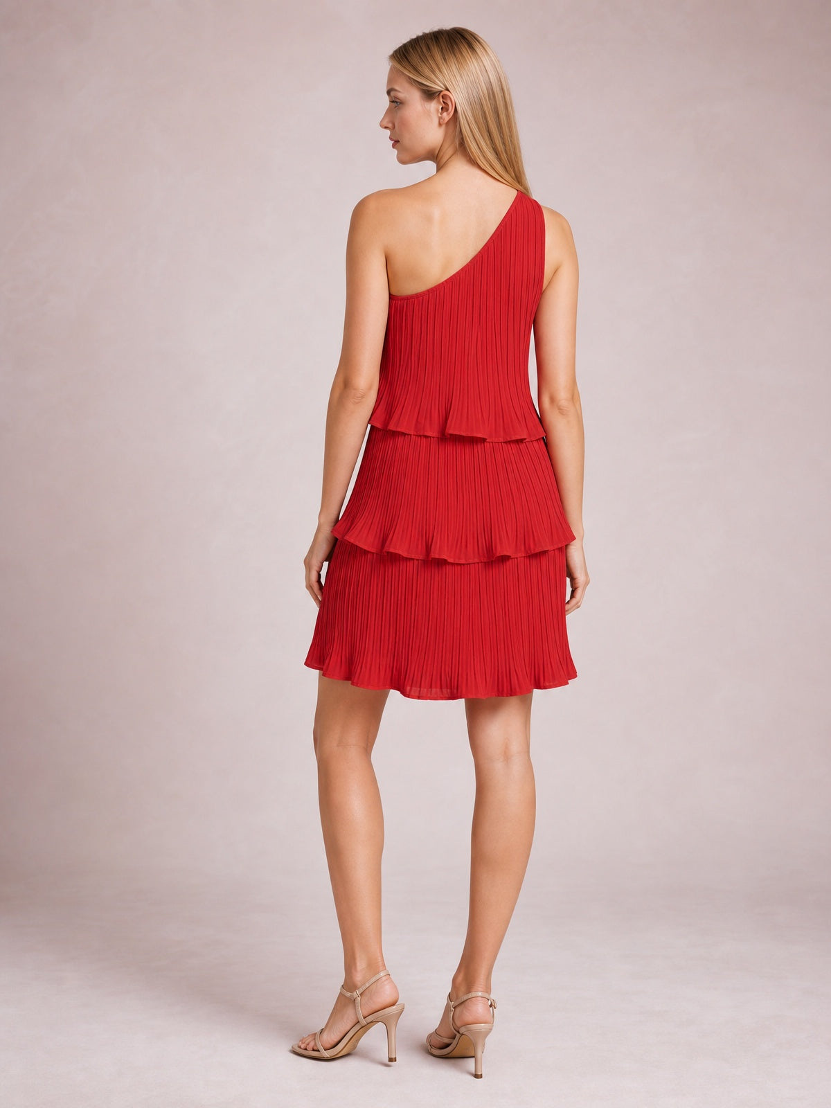 A model stands in a red layered single-shoulder mini dress, shown from behind with a pleated texture, tiered ruffled silhouette, and asymmetrical neckline.