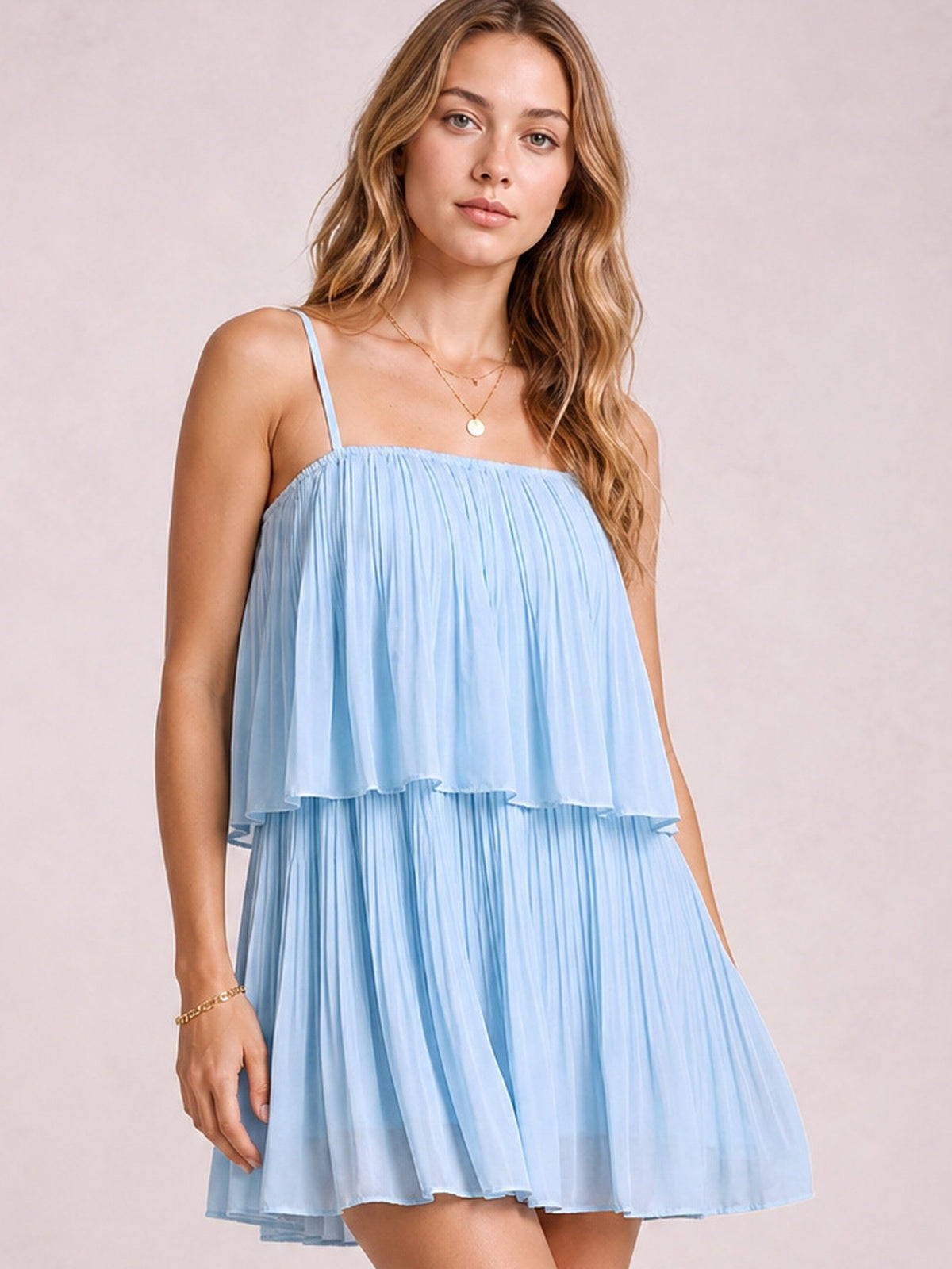 A model wears a sky blue layered mini cami dress with delicate straps, a softly gathered square neckline, ruffled overlay, and flowing pleated silhouette.
