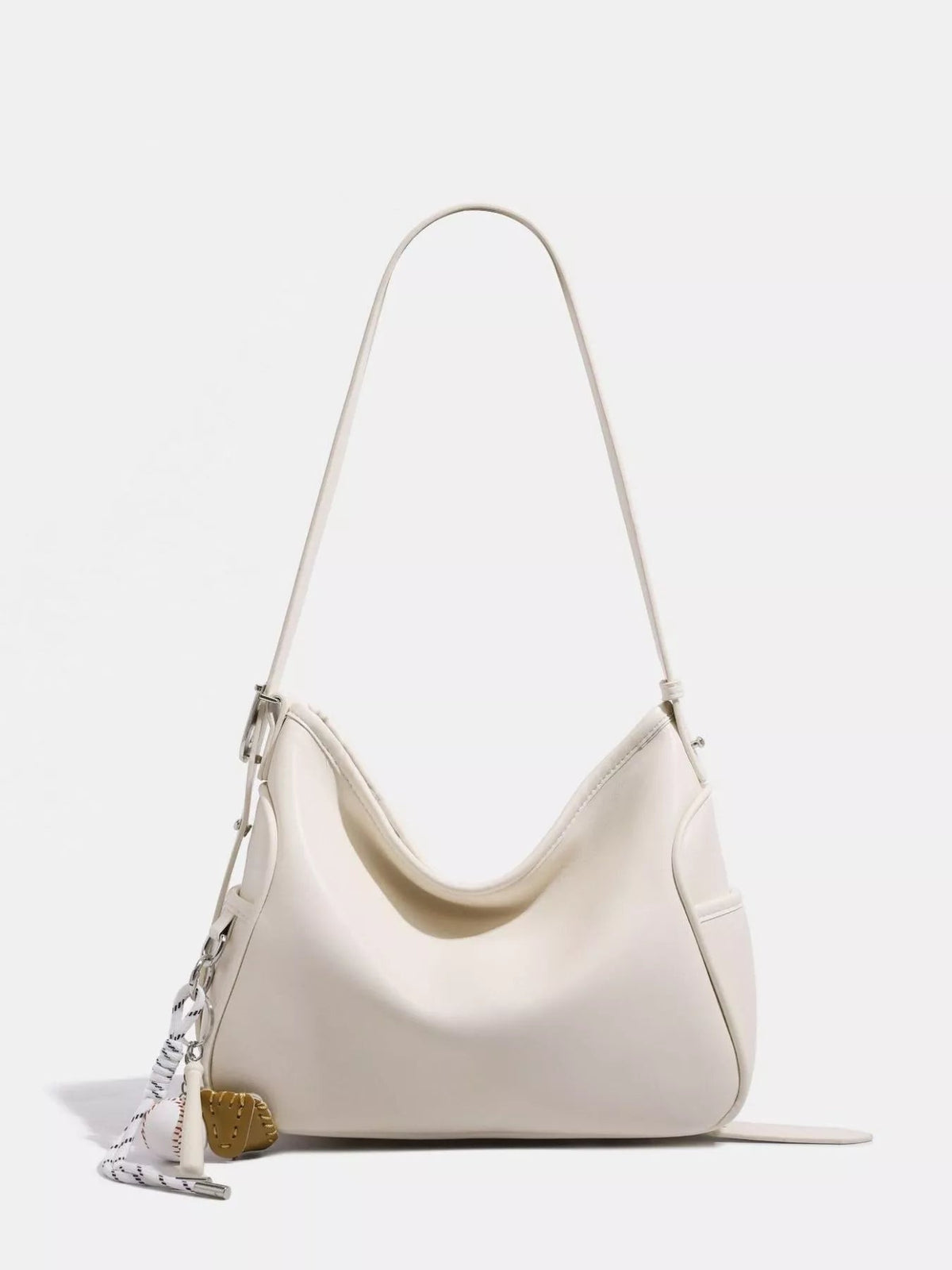 Cream PU leather slouchy shoulder bag with a medium-size curved silhouette, long adjustable strap, softly gathered top, side hardware, and decorative charms.