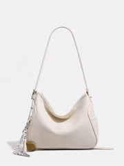 Cream PU leather slouchy shoulder bag with a medium-size curved silhouette, long adjustable strap, softly gathered top, side hardware, and decorative charms.