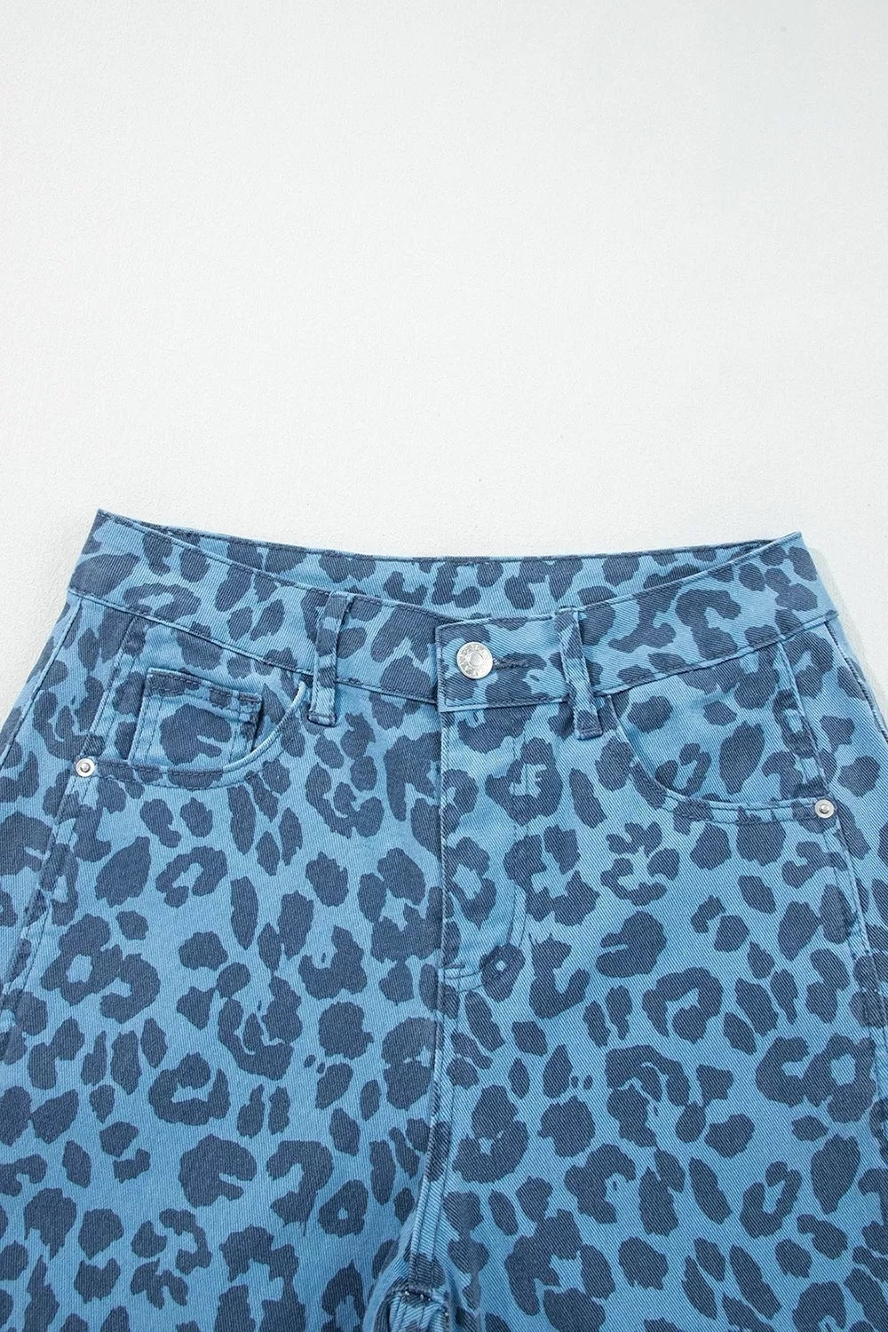 Blue leopard print jeans shown in close-up feature a structured waistband, belt loops, front button closure, angled pockets, and contrast dark spots.