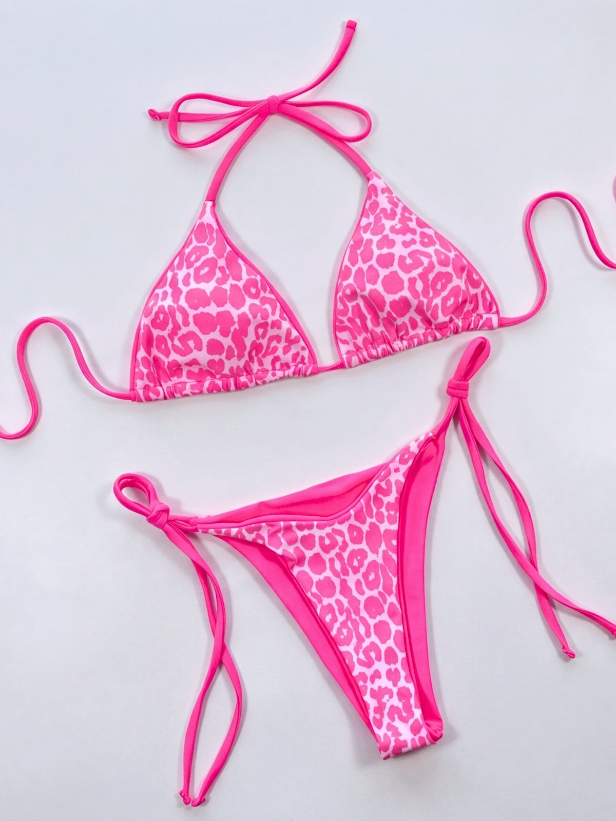 Pink-and-white leopard print bikini set laid flat, featuring a triangle halter top and matching tie-side bottoms with bright pink trim and straps.