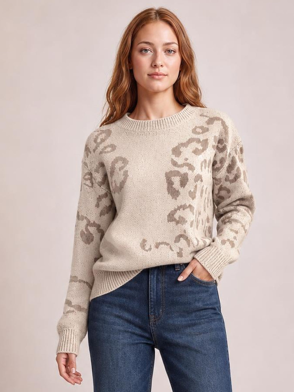 Model poses with one hand in her jeans pocket wearing a beige relaxed fit leopard sweater with a crew neckline, long sleeves, and ribbed trim.