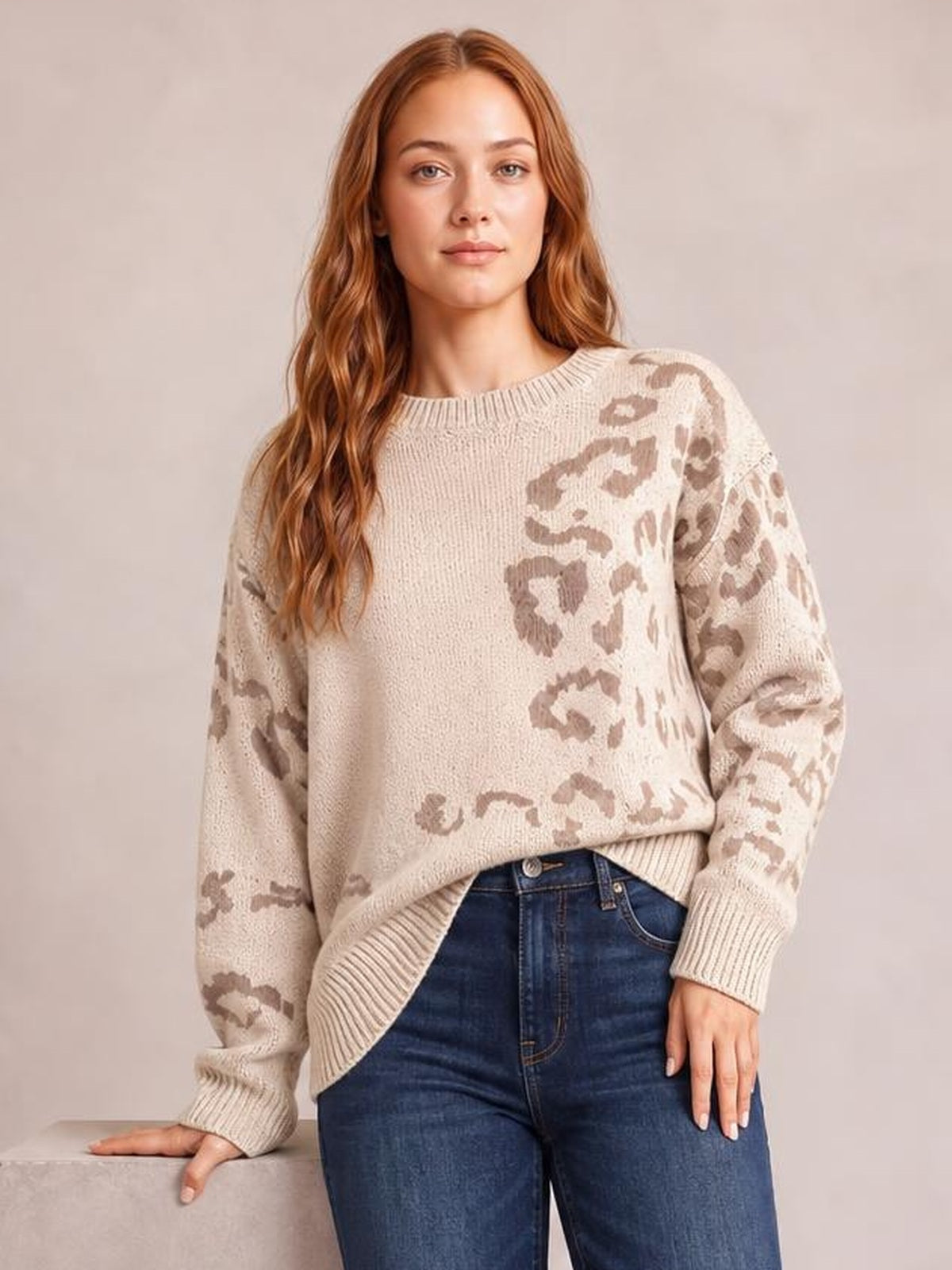 A model poses in a relaxed-fit beige leopard-print sweater with a ribbed crew neckline, cuffs, and hem, resting one hand on a ledge.