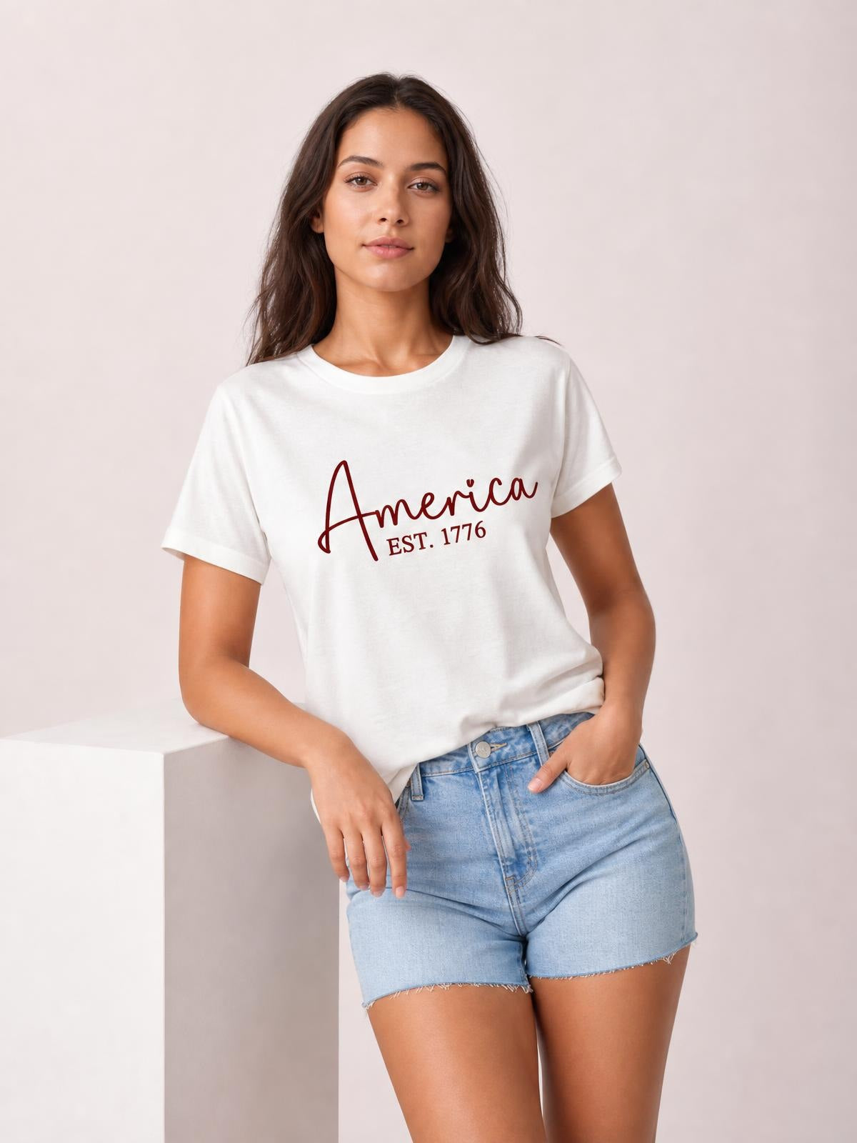 Model wears a white round neck short sleeve T-shirt with red “America EST. 1776” letter graphic, relaxed silhouette, and ribbed neckline.