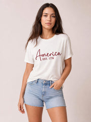 A model poses in a white round-neck short-sleeve T-shirt with rolled cuffs, a red “America EST. 1776” letter graphic, and a relaxed silhouette.