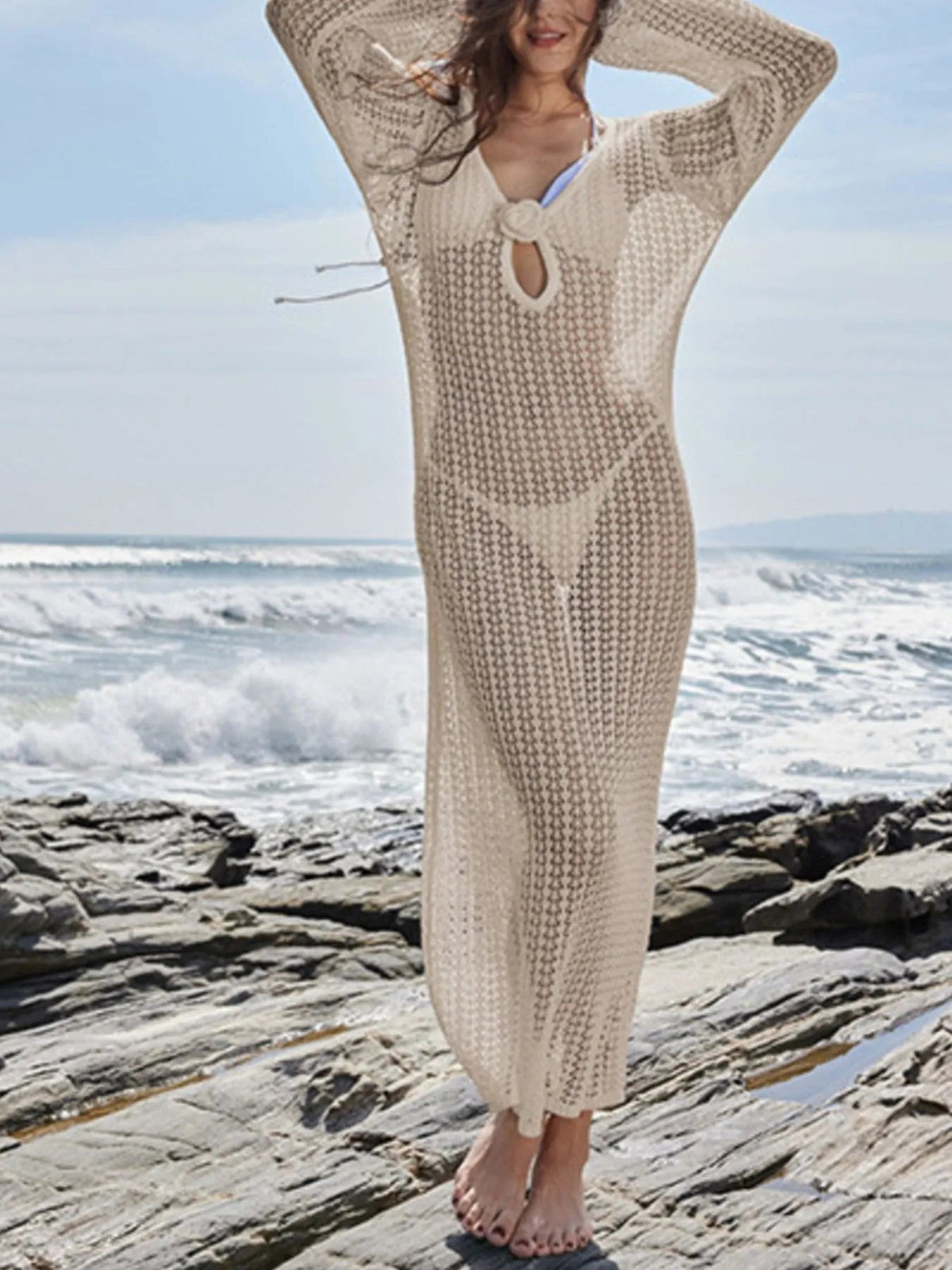 Model wears a cream long-sleeve open-knit cover-up dress with a slim ankle-length silhouette, tied front detail, and arms raised overhead.
