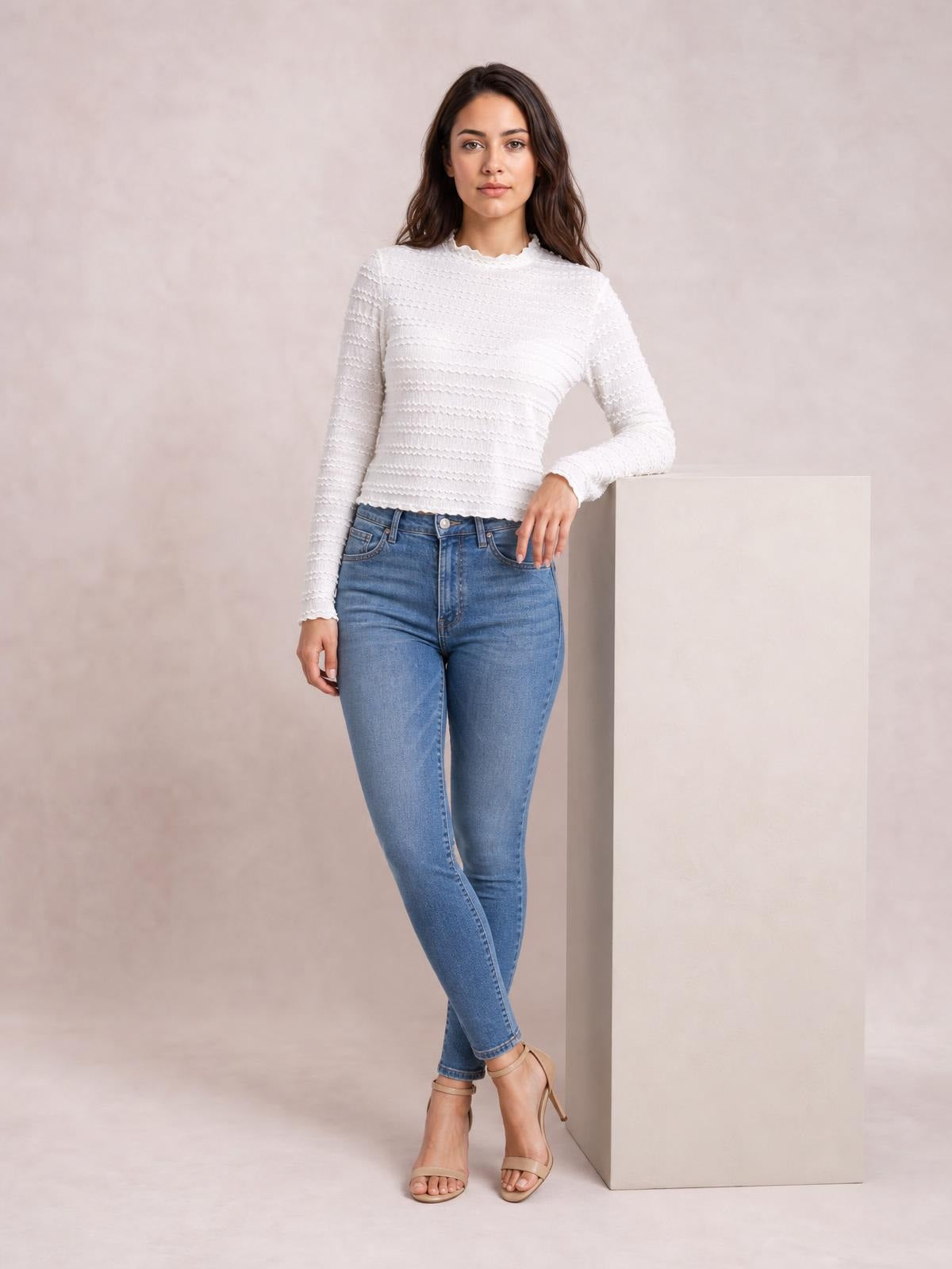 Model wears a white long-sleeve jacquard slim top with textured horizontal detailing and a fitted silhouette, leaning against a pedestal with crossed legs.