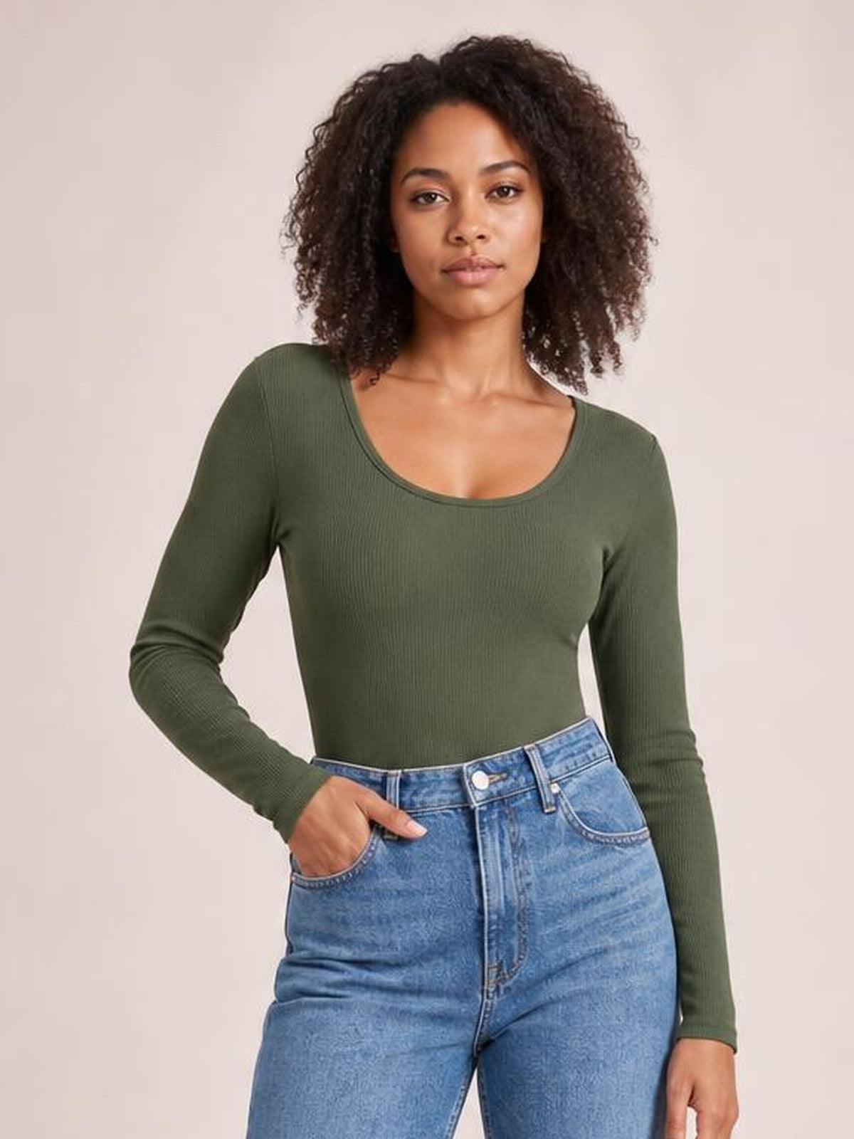 Model poses with one hand in her jeans pocket wearing an olive green long sleeve scoop neck T-shirt with a fitted silhouette and stretch construction.
