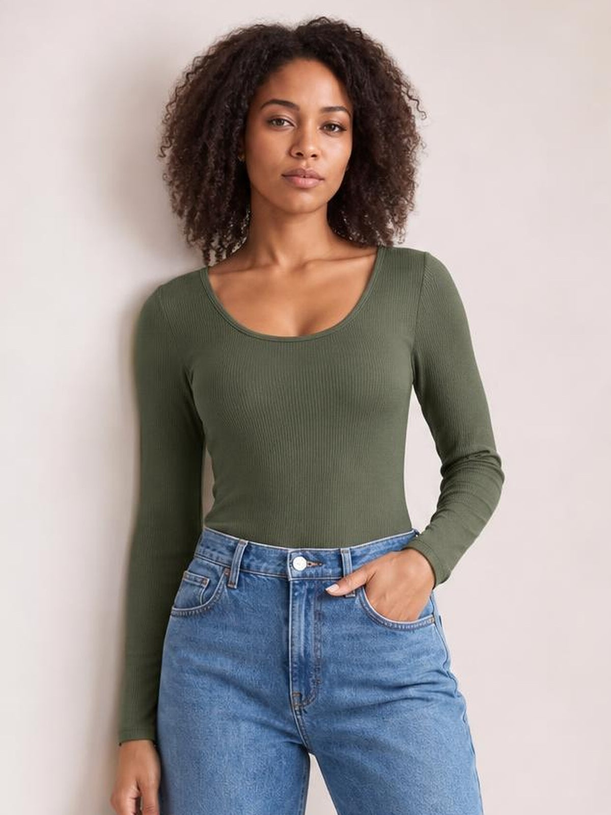 Model wears an olive green long sleeve scoop neck T-shirt with a fitted silhouette and ribbed construction, posing with one hand in her jeans pocket.