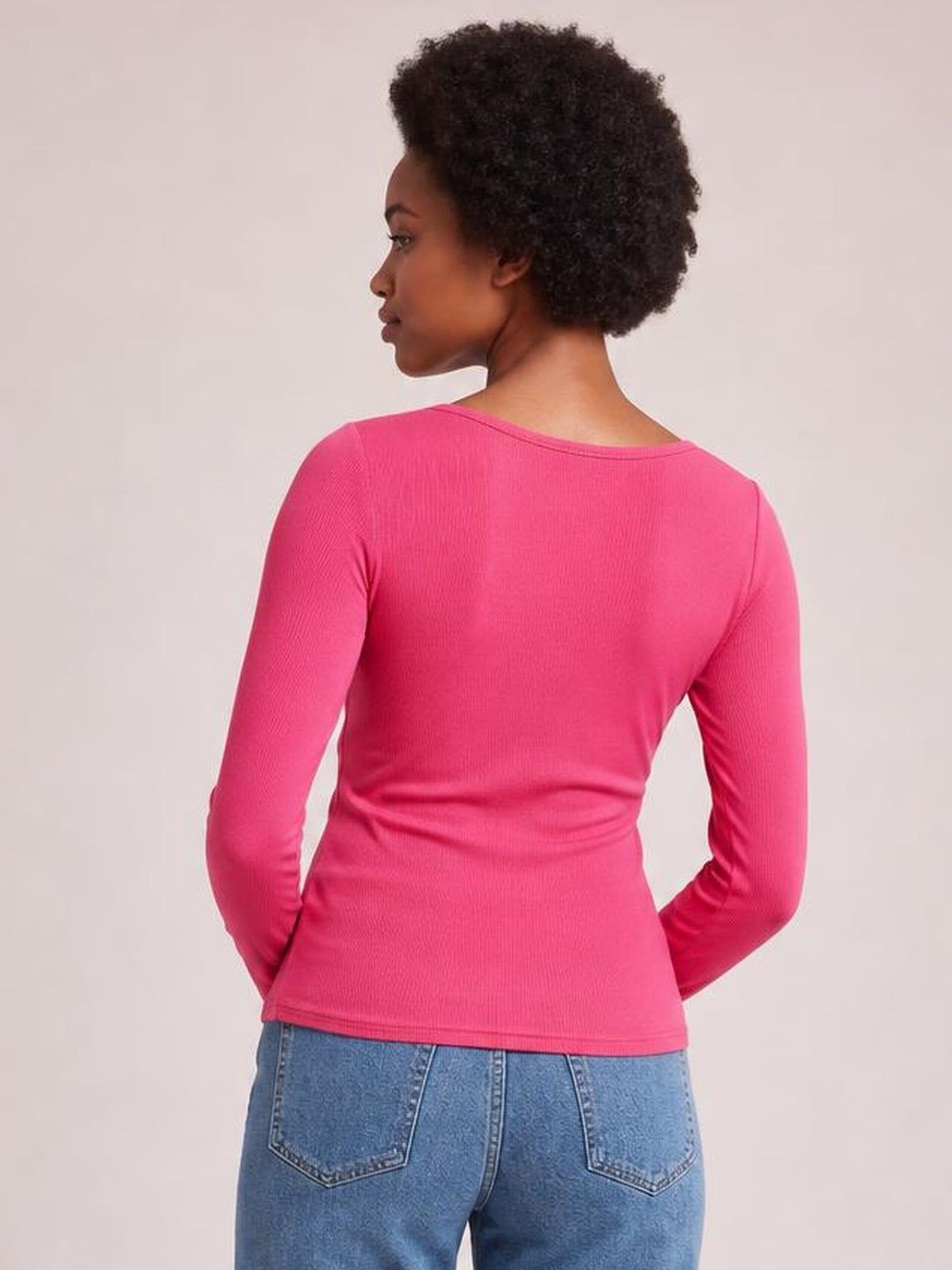 A model poses with her hands behind her in a bright pink long sleeve scoop neck T-shirt featuring a fitted silhouette, fine ribbed texture, and straight hem.