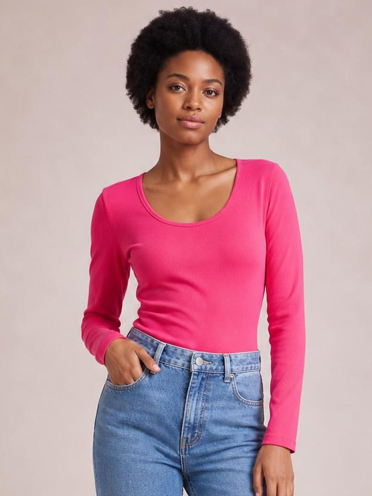 Model wearing a bright pink long sleeve scoop neck T-shirt with a fitted silhouette and stretch construction, posing with one hand in her jeans pocket.