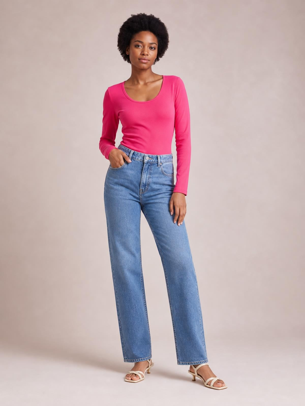 Model poses in a bright pink long sleeve scoop neck T-shirt with a fitted silhouette, ribbed construction, and one hand tucked into her jeans pocket.