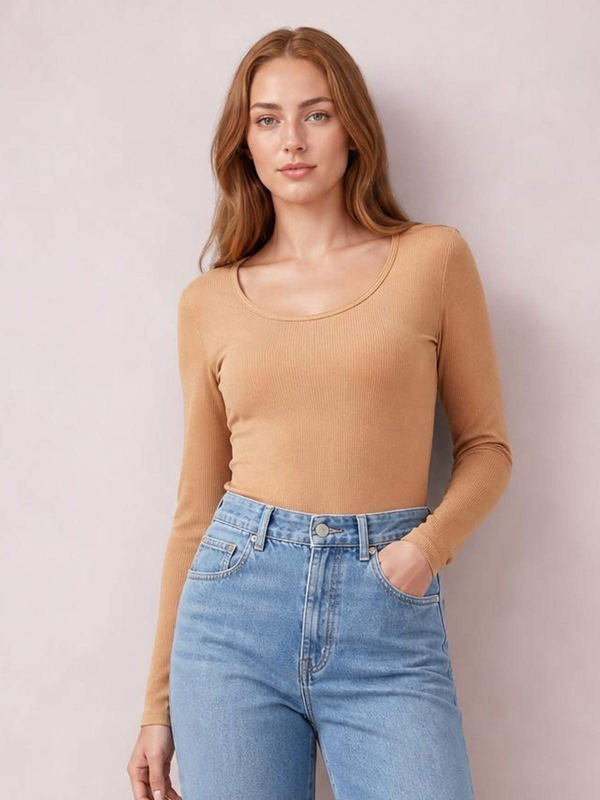 A model wears a tan long sleeve scoop neck T-shirt with a fitted silhouette, fine textured construction, and one hand tucked into her jeans pocket.