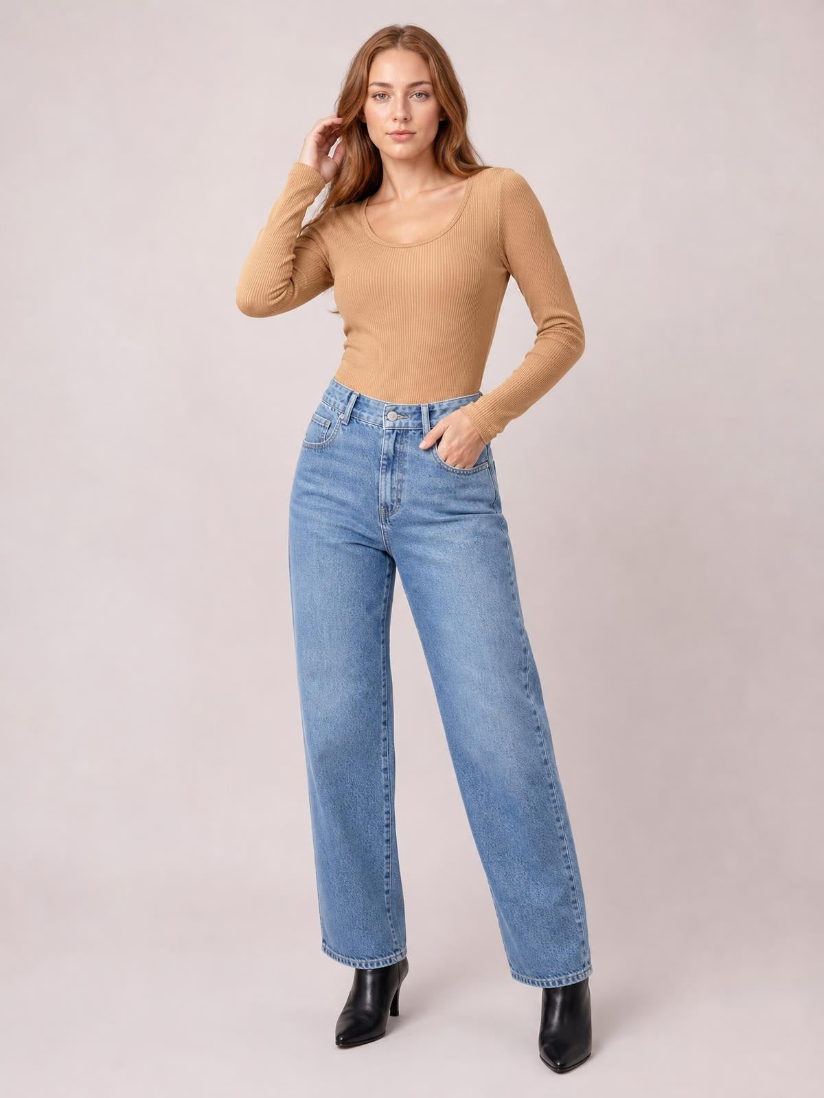 A woman models a camel long sleeve scoop neck T-shirt with a fitted ribbed silhouette, posing with one hand in her jeans pocket and the other touching her hair.