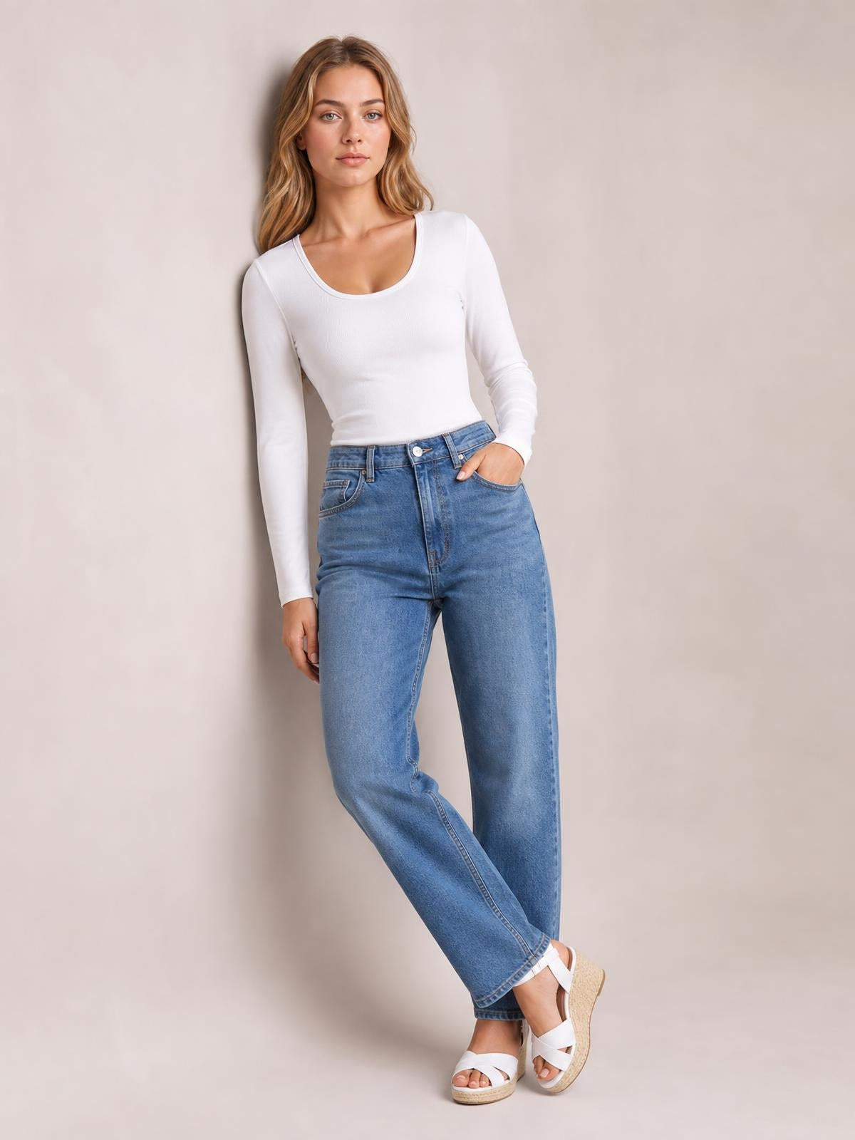 Model wears a white long sleeve scoop neck T-shirt with a fitted silhouette and stretch construction, posing with one hand in her jeans pocket.