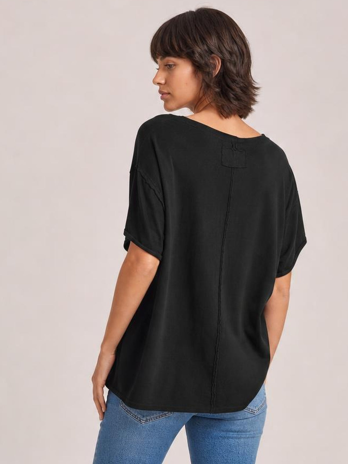Model wearing a black exposed seam round neck half sleeve T-shirt with a relaxed, loose silhouette and visible center-back and shoulder seam details.