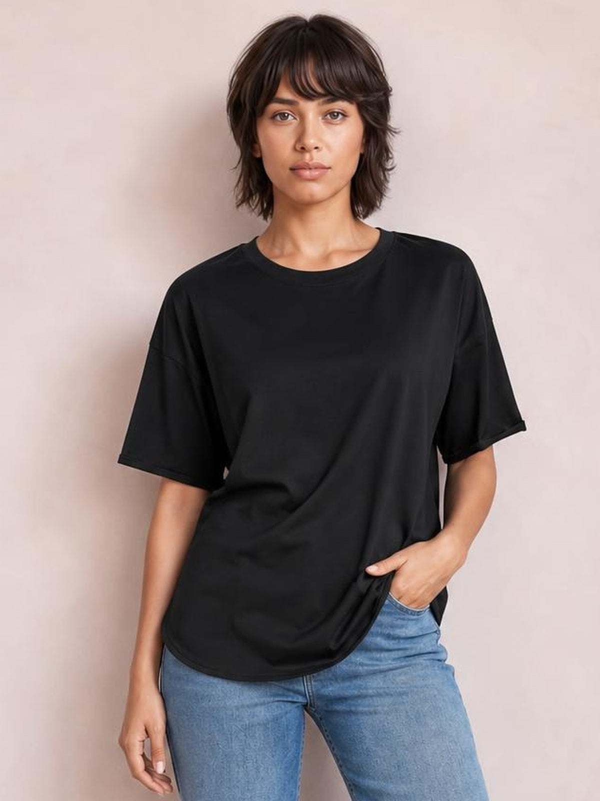A model wears a black round-neck half-sleeve T-shirt with a relaxed silhouette and subtle exposed seam construction, posing with one hand in her jeans pocket.
