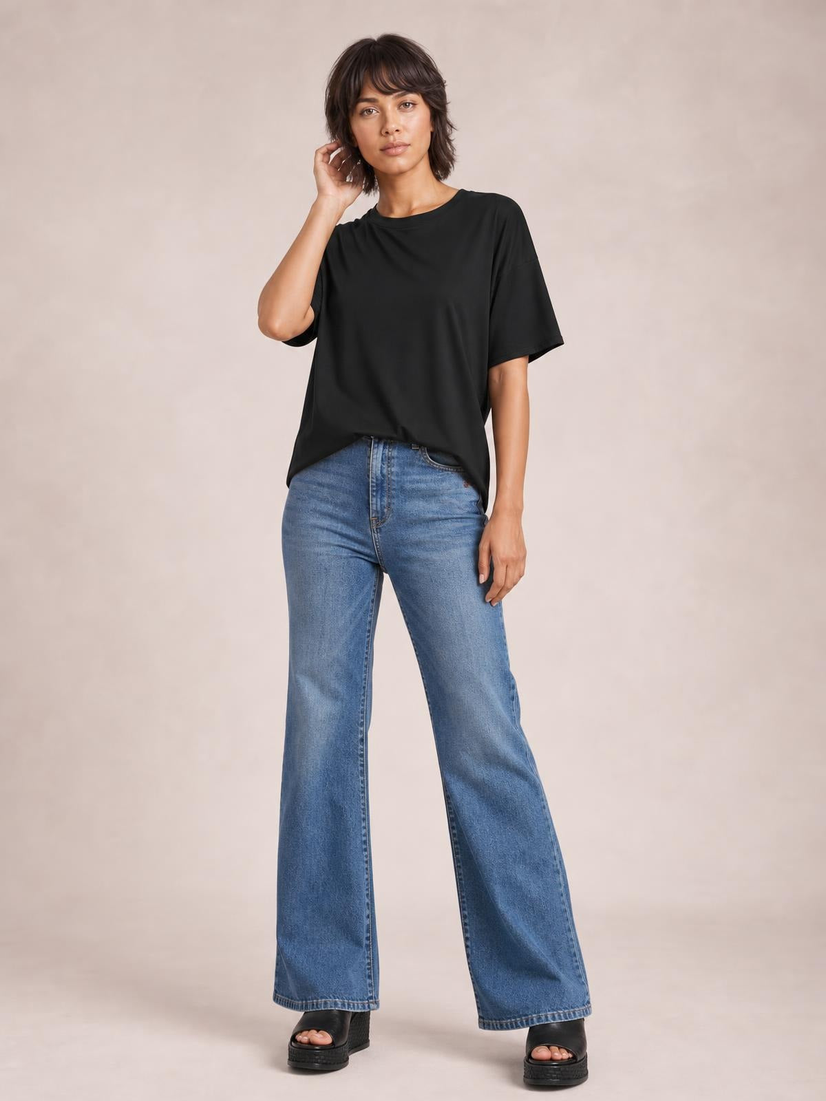 Model wears a black round-neck half-sleeve T-shirt with a relaxed silhouette and exposed seam construction, posing with one hand near her hair.