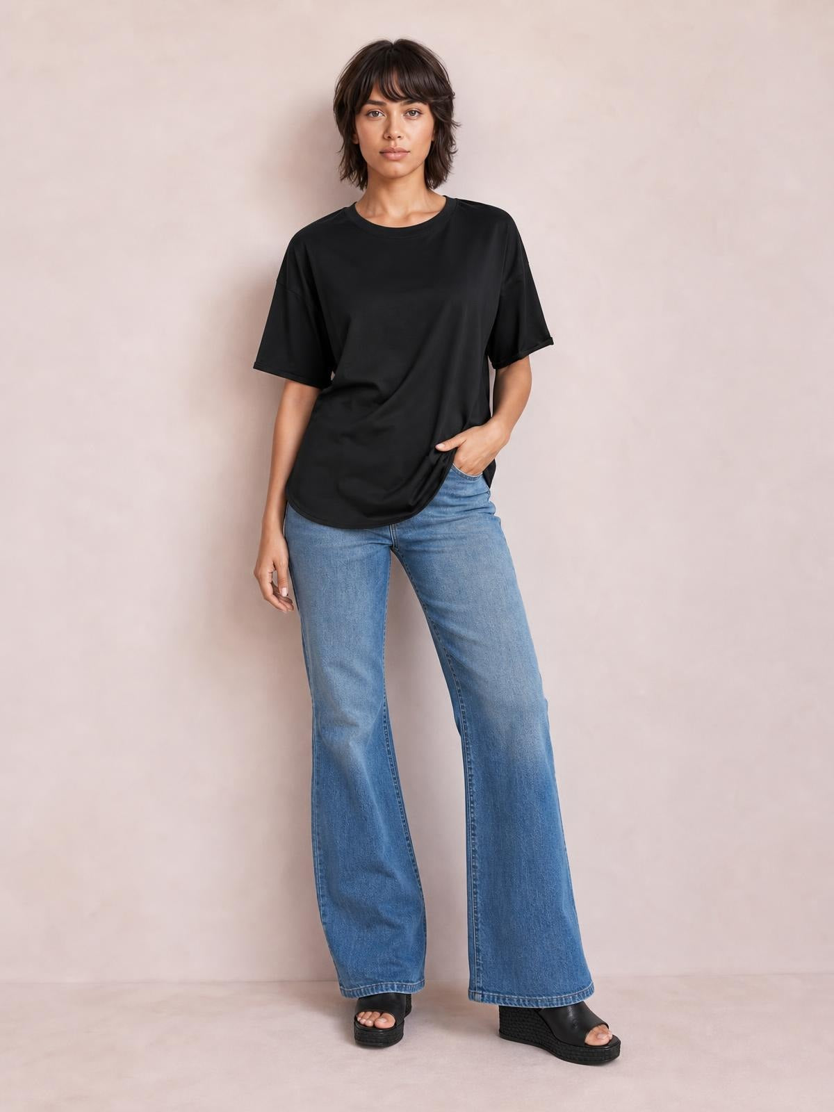 Model wears a black exposed seam round neck half sleeve T-shirt with a relaxed silhouette, curved hem, and one hand tucked into her jeans pocket.
