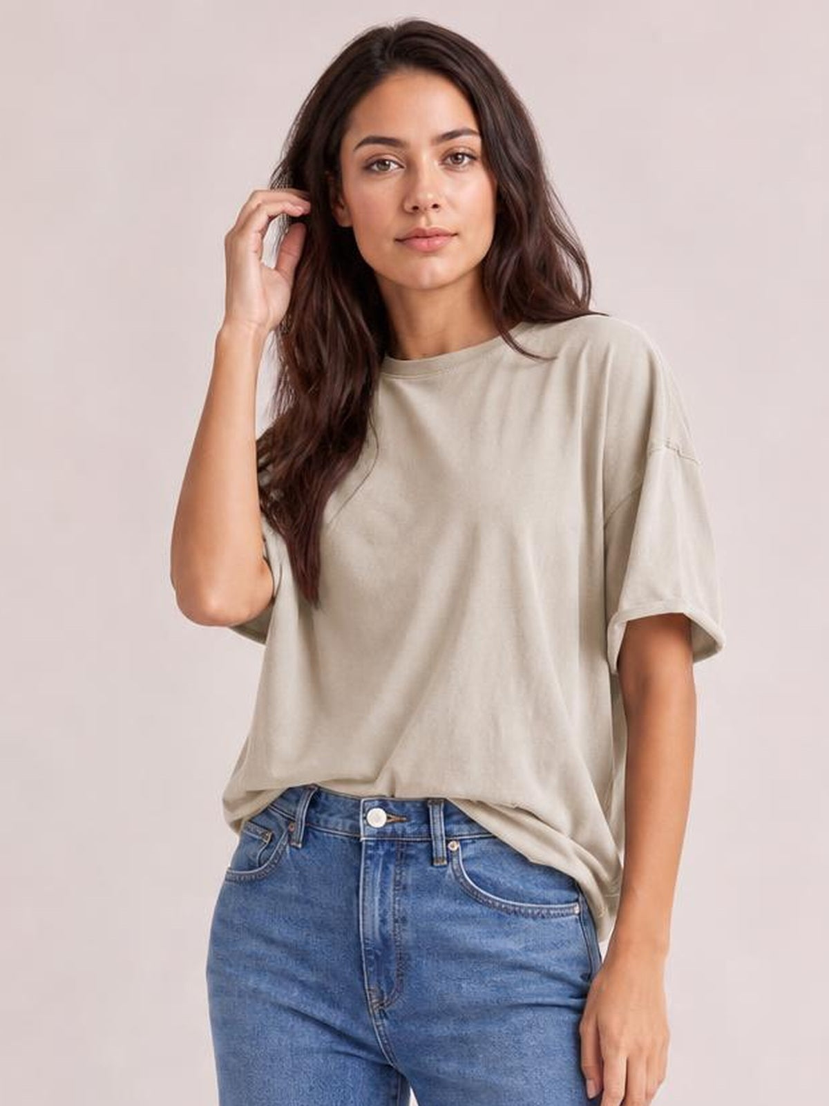 Model wearing a beige exposed seam round neck half sleeve T-shirt with a relaxed silhouette, paired with blue jeans and one hand touching her hair.