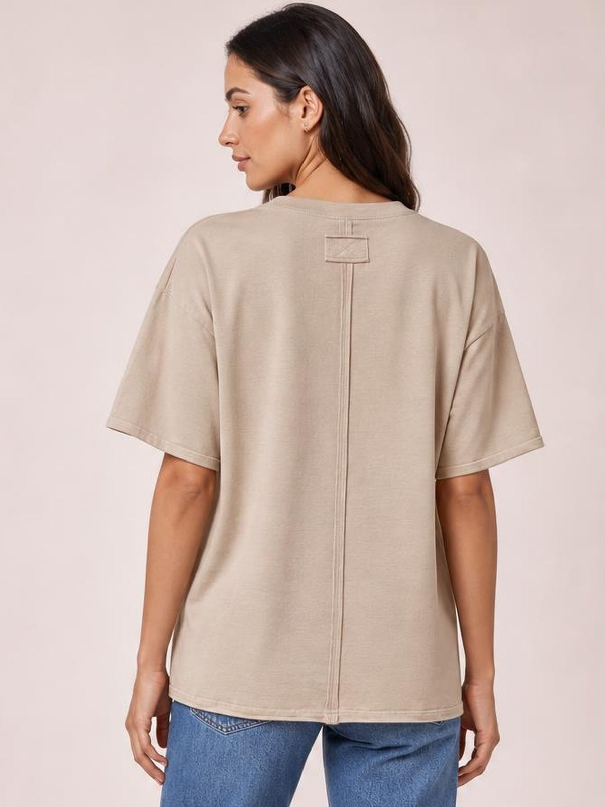 A model seen from behind wears a beige round-neck half-sleeve T-shirt with exposed center-back seams, a stitched patch, and a relaxed straight silhouette.