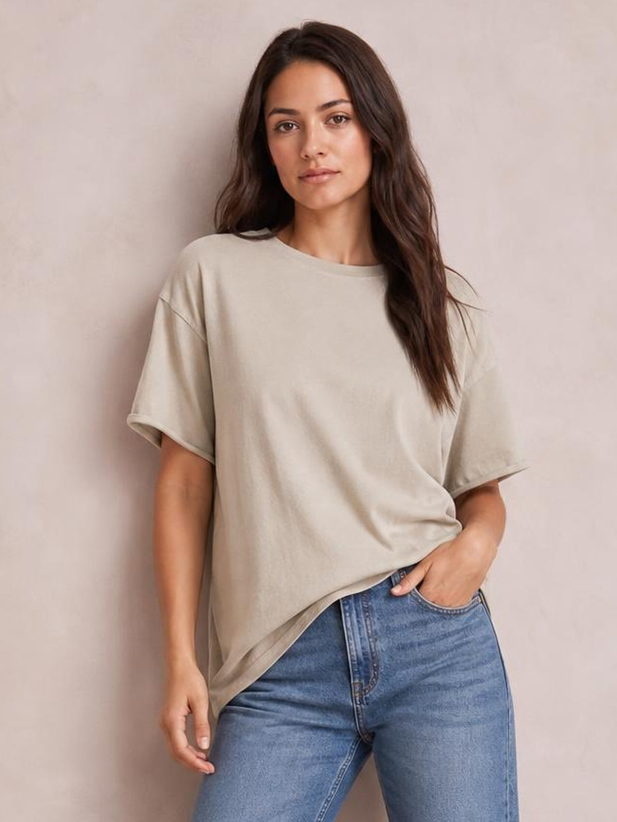 Model wears a taupe exposed-seam round-neck half-sleeve T-shirt with a relaxed silhouette, draped hem, and one hand resting in her jeans pocket.