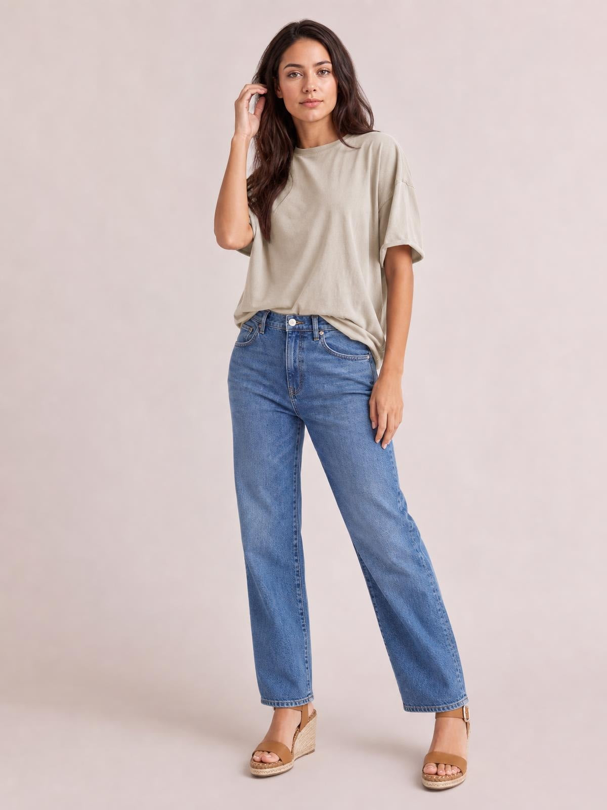 Model wears a taupe exposed seam round neck half sleeve T-shirt with a relaxed, slightly oversized silhouette, posing with one hand near her hair.